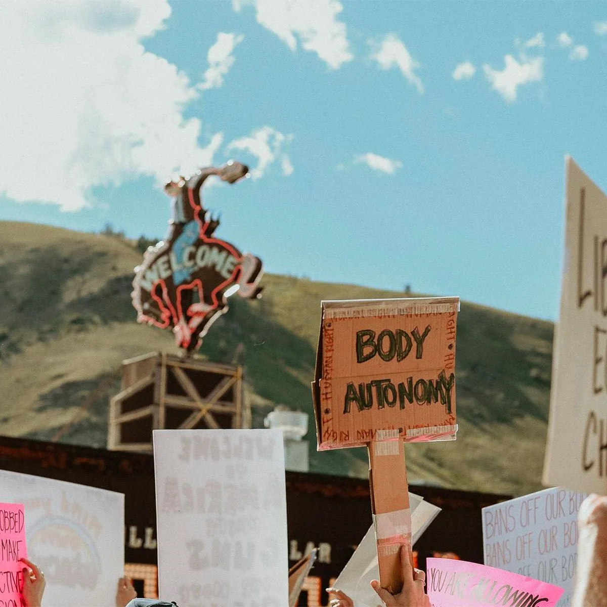 Protesters at a demonstration holding signs, one reading 'Body Autonomy,' with a hill and a large neon sign in the background under a partly cloudy sky.