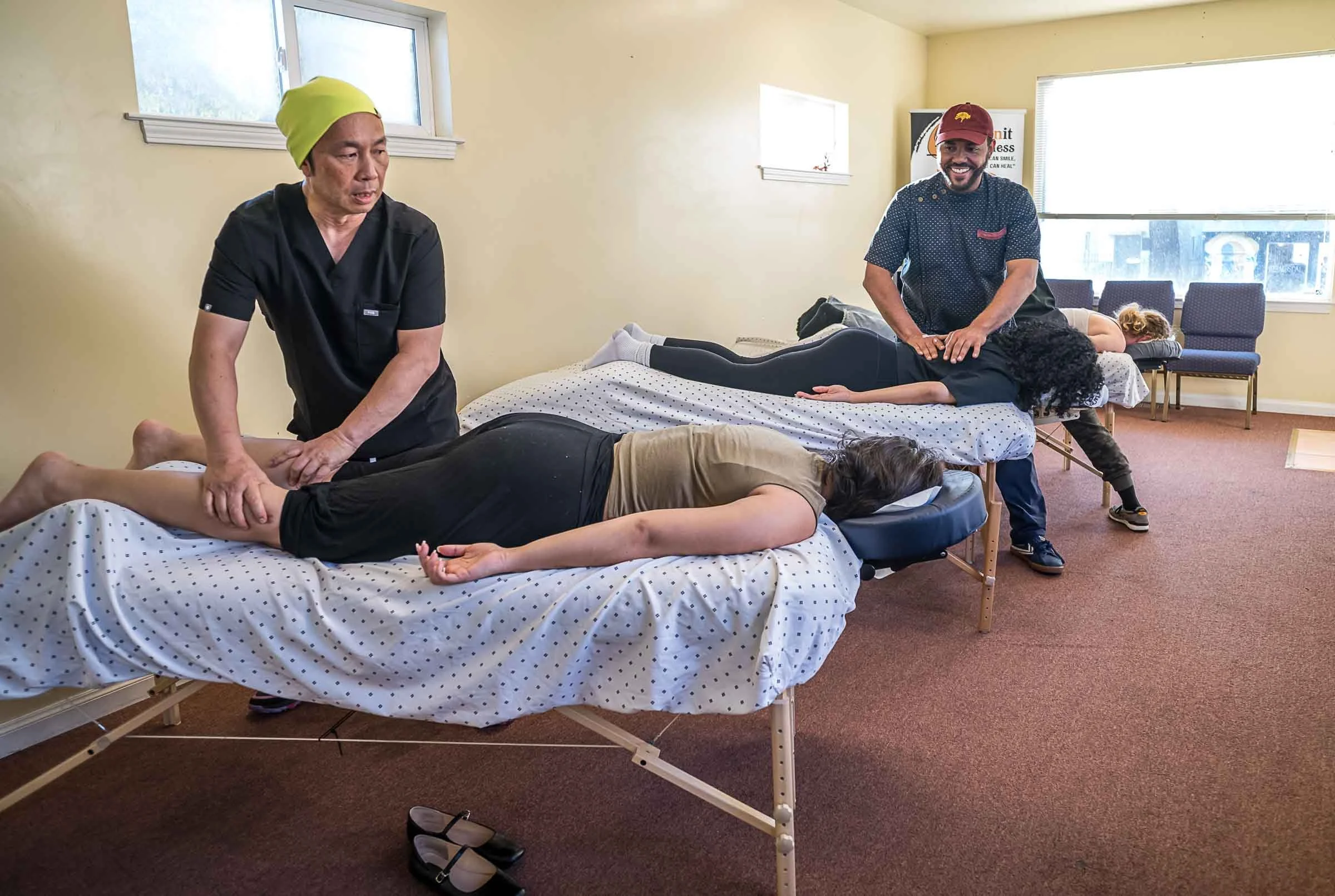 Two people are lying face down on beds, receiving massage therapy from two male massage therapists. The room has a yellow wall, a window, and several chairs against the wall, with one person sitting on a chair. The scene appears to be in a wellness or massage clinic.