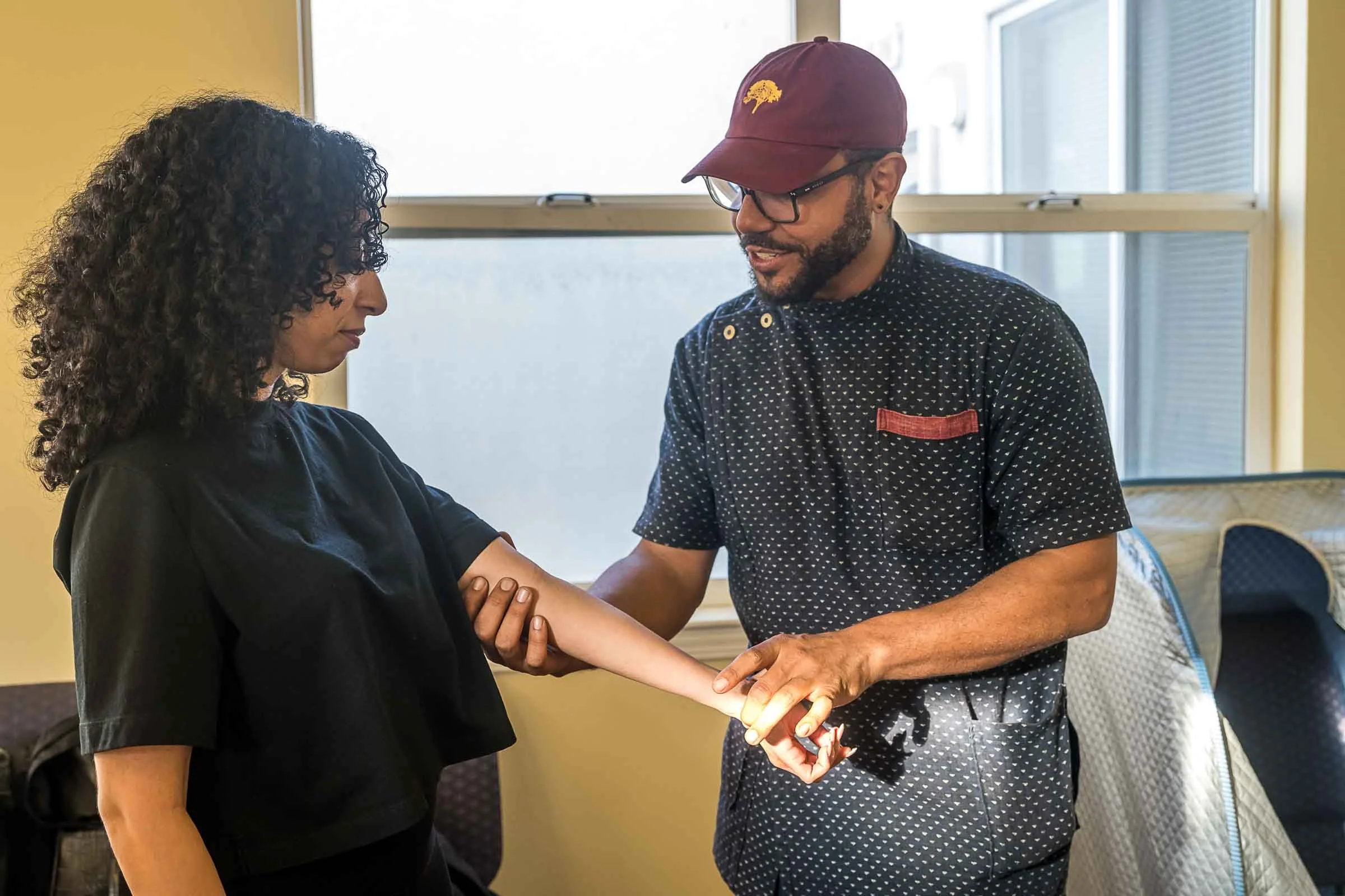 A man and woman sharing a moment, holding hands and looking at each other. The woman has curly dark hair and is dressed in a black shirt. The man is wearing glasses, a maroon cap with a yellow tree logo, and a black shirt with white dots. They are indoors near a window, smiling softly.