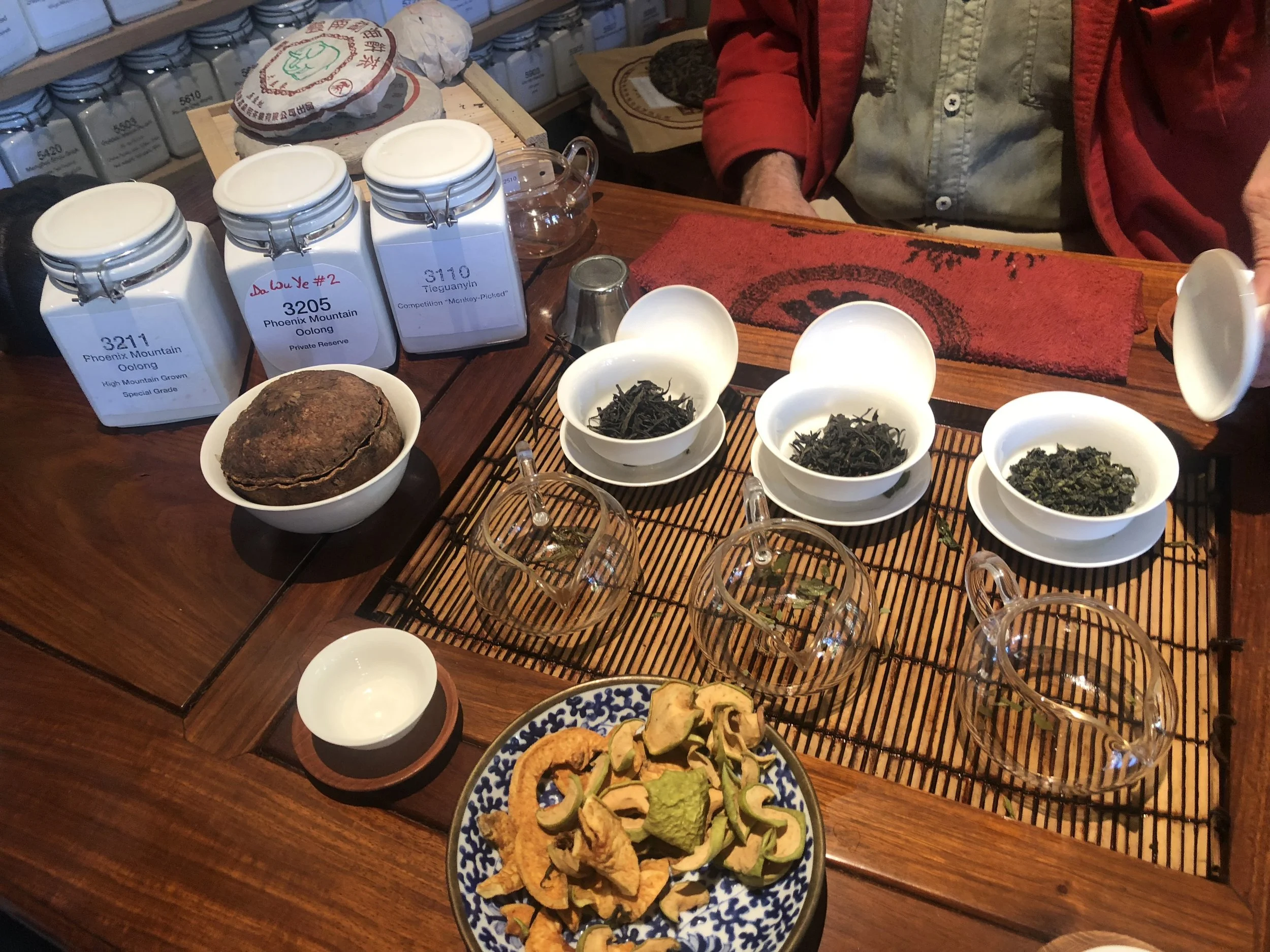 Various teas and snacks arranged on a wooden table, including jars labeled with tea types, bowls of loose tea leaves, a round pressed tea cake, and a bowl of mixed dried vegetables or herbs, with a person in a red jacket and beige shirt partially visible in the background.