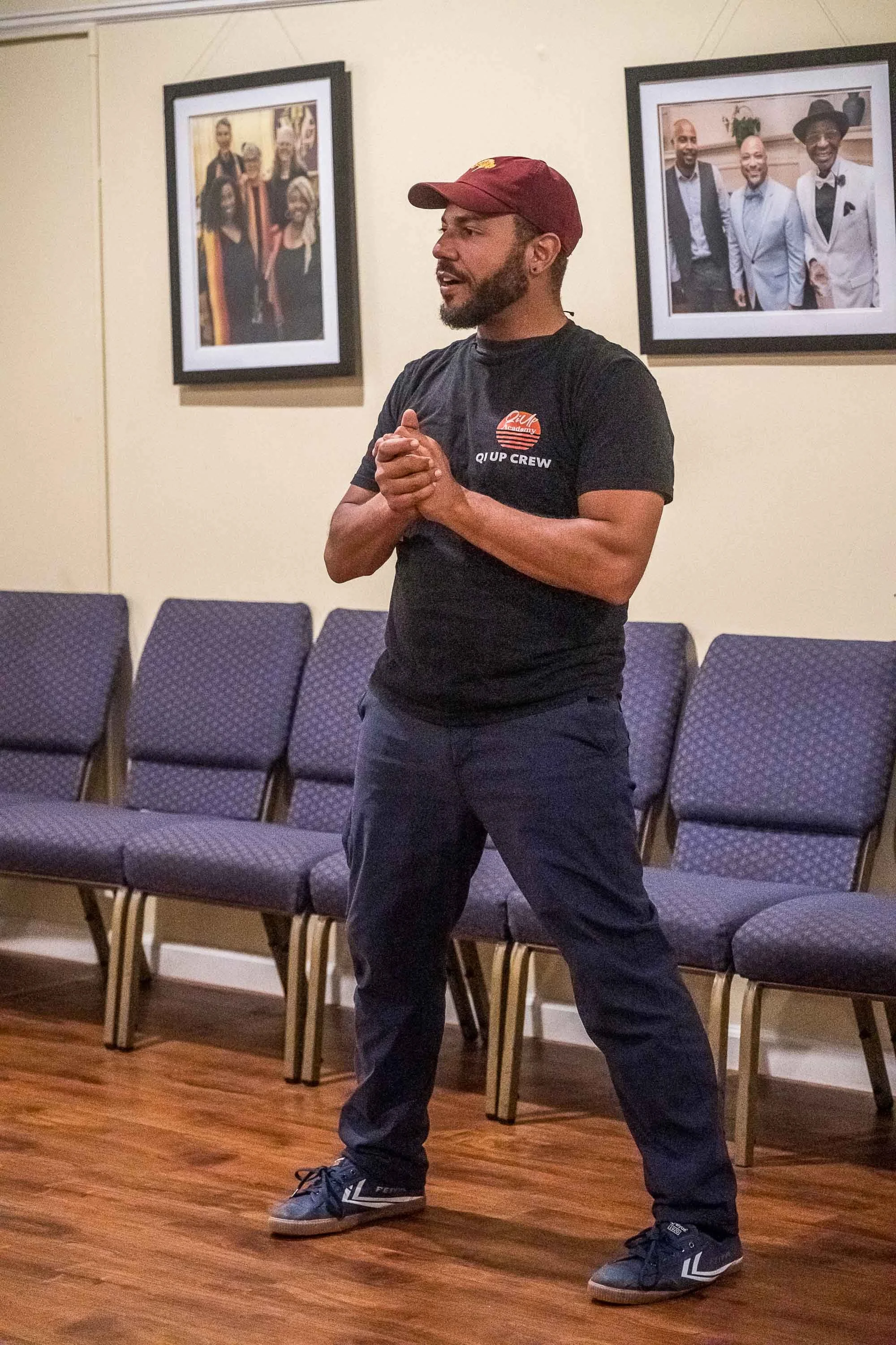 A man wearing a maroon cap, black t-shirt, dark pants and sneakers is speaking or presenting in an indoor space with a row of empty chairs behind him and framed photos on the wall.