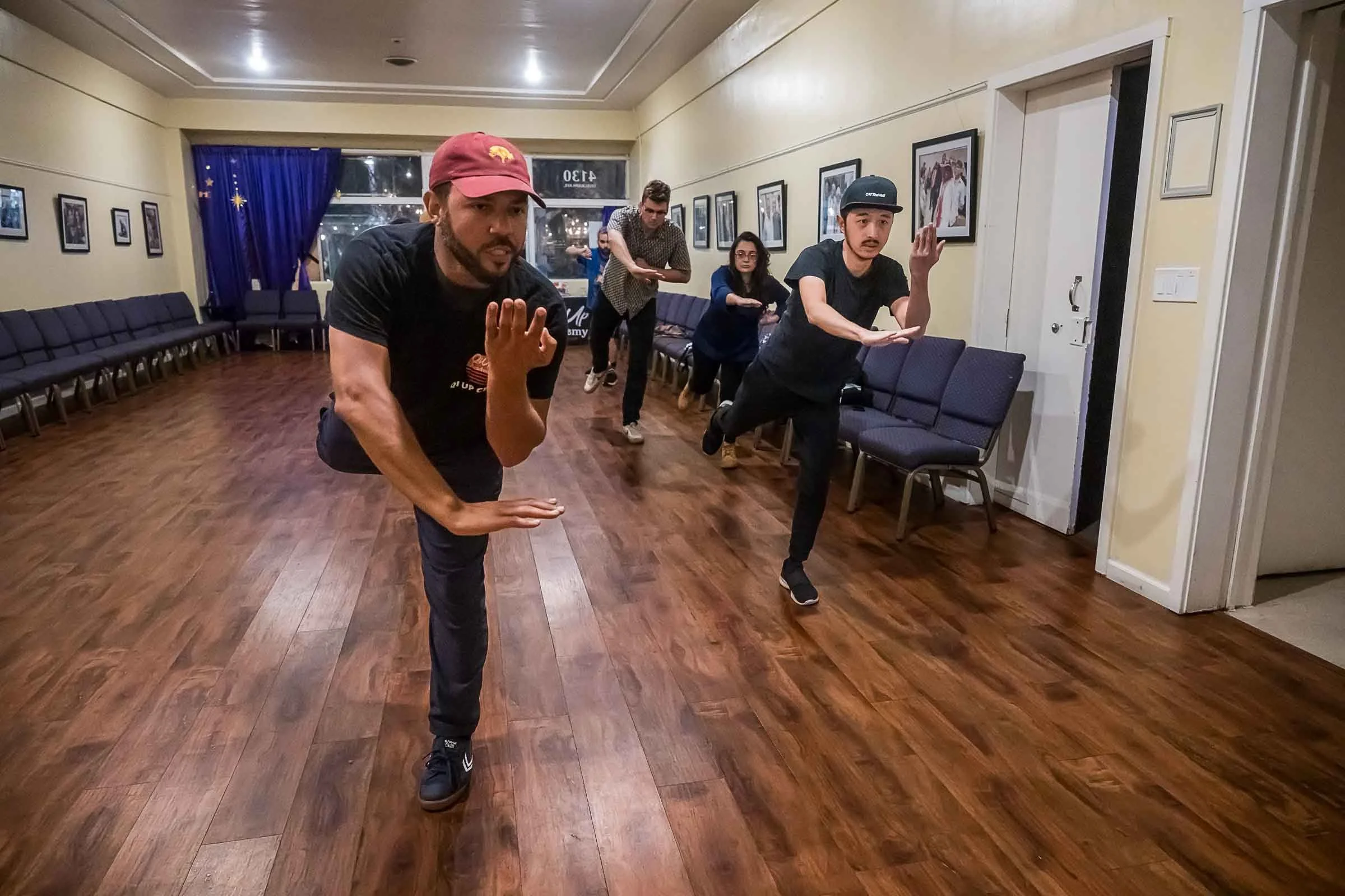 People engaging in a dance or martial arts practice in a room with wood floors and framed photos on the walls, some of the individuals are wearing hats and casual clothing.