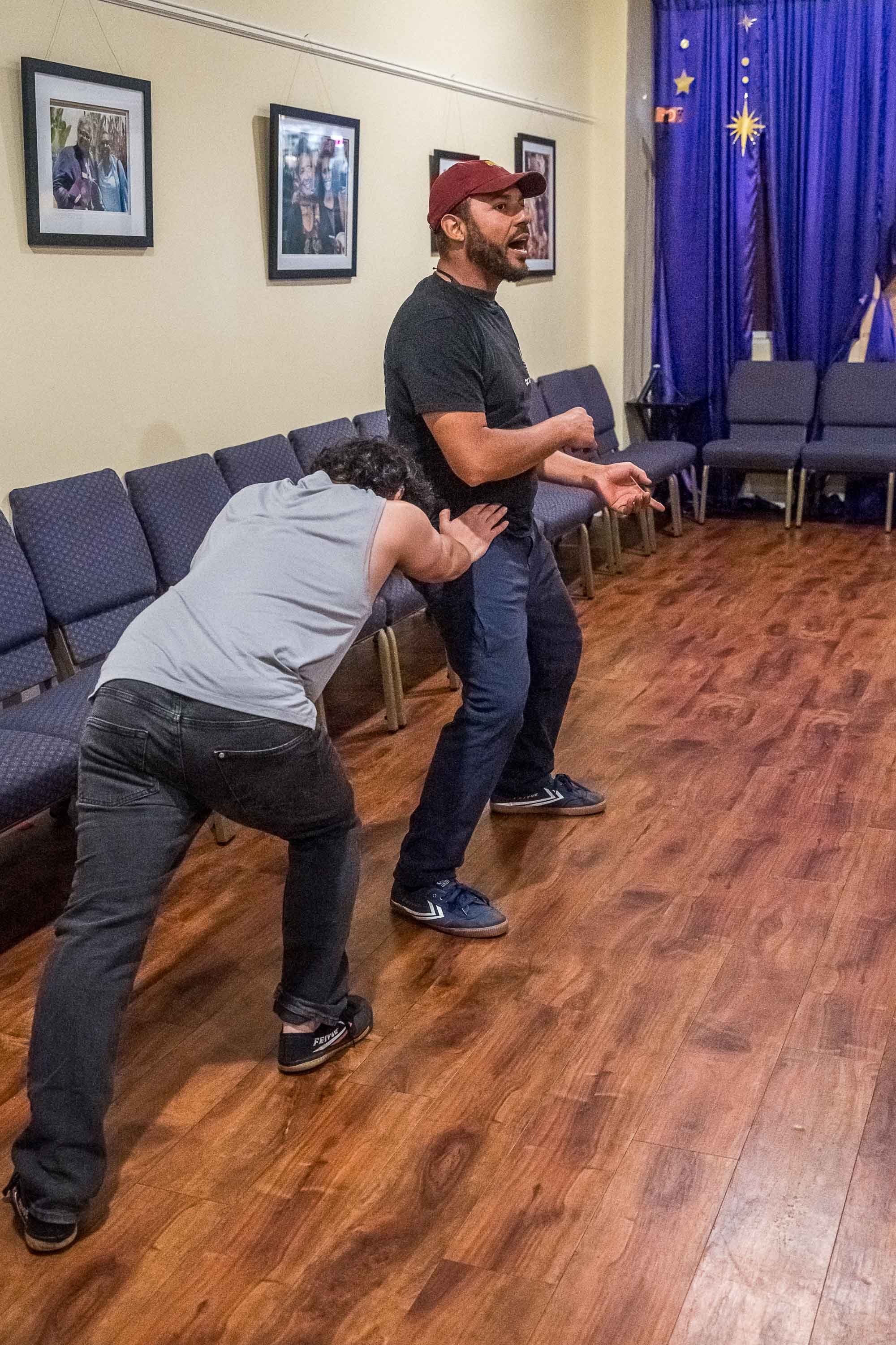A man wearing a red cap and black shirt appears to be singing or speaking passionately while a woman in a gray tank top and black pants appears to be either dancing or falling, with her head leaning on the man's waist. They are in a room with framed pictures on the wall, wooden floor, and navy blue curtains decorated with stars.