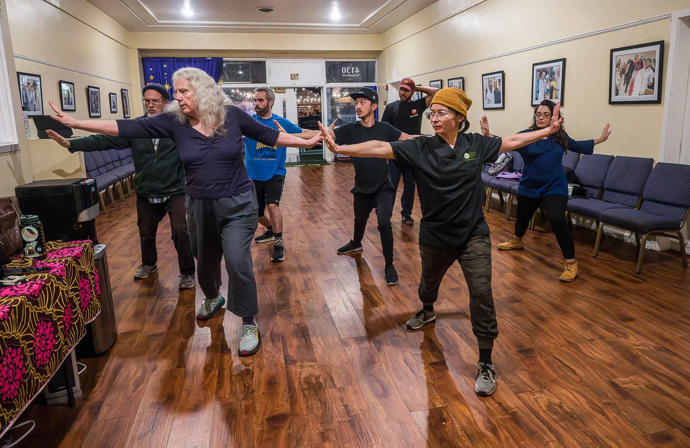 Group of diverse people practicing a dance or yoga routine in a room with framed photographs on the walls, wooden floor, and casual attire.