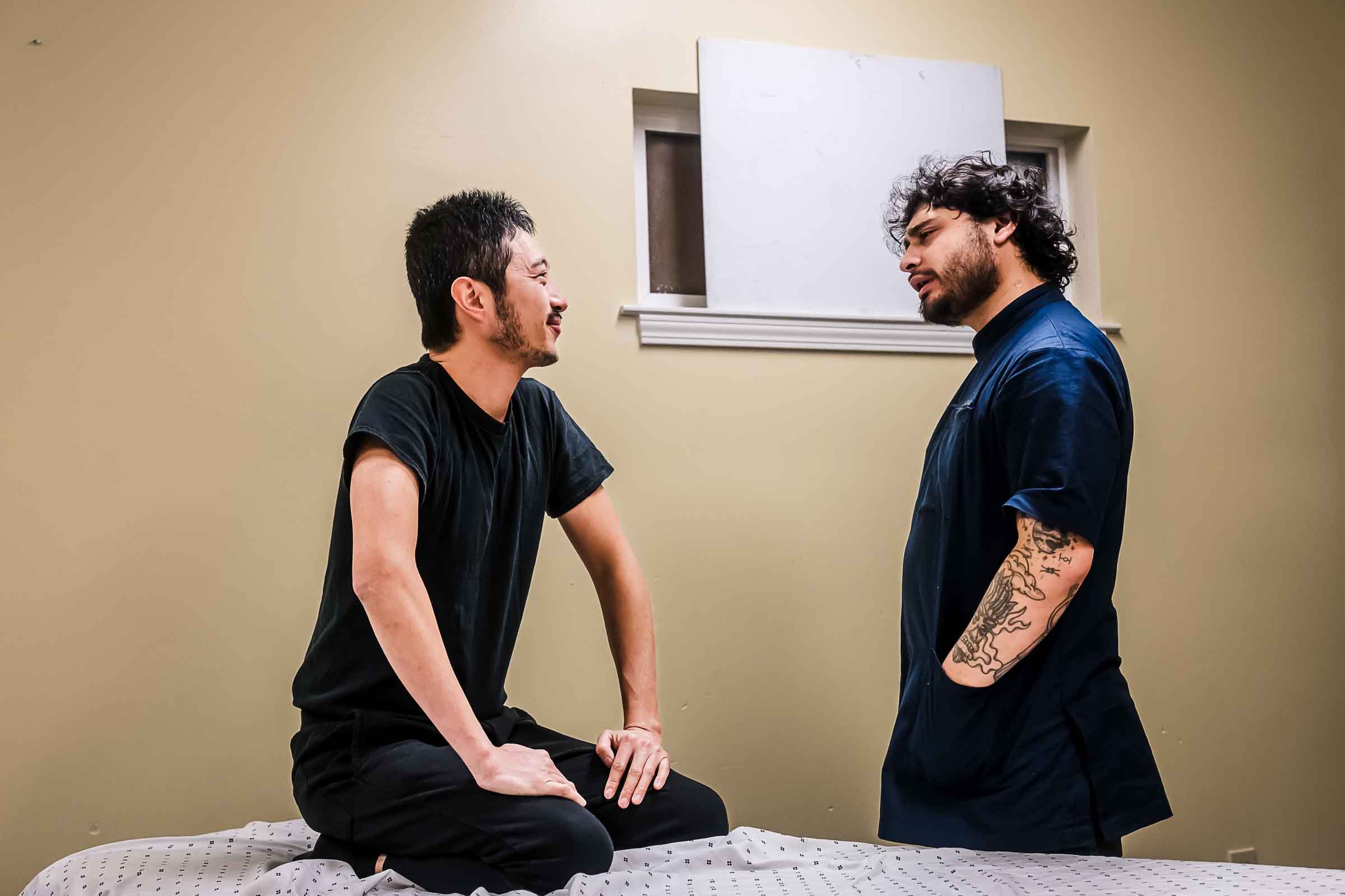 Two men engaged in a serious conversation in a room with beige walls, one sitting on a bed and the other standing, with a window behind them.