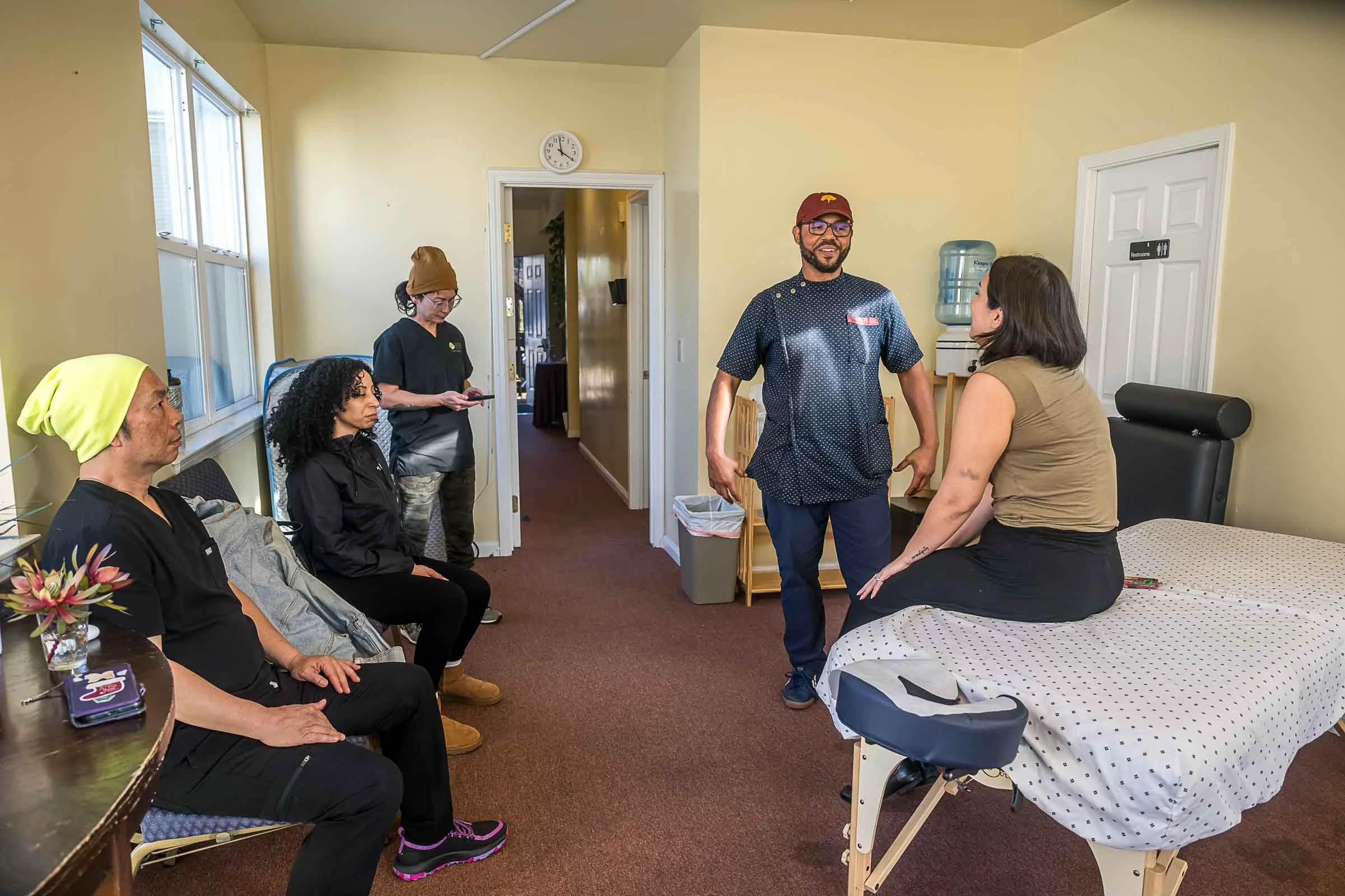 A group of five people in a hospital room, some sitting on chairs and one standing, engaged in conversation. The room has yellow walls, a water cooler, a closed restroom door, and a bed with a patient. The group appears to be discussing or having a consultation.