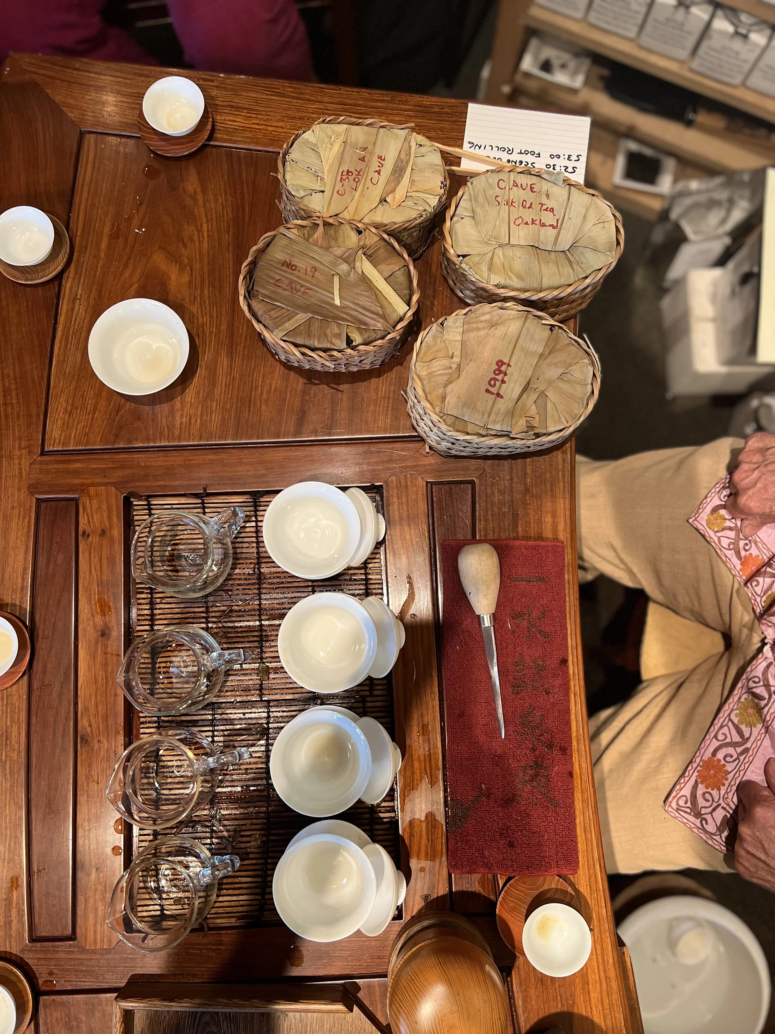 Empty cups and glasses placed on a bamboo mat on a wooden table in a shop or café, with woven baskets containing dried leaves and handwritten notes on the table.