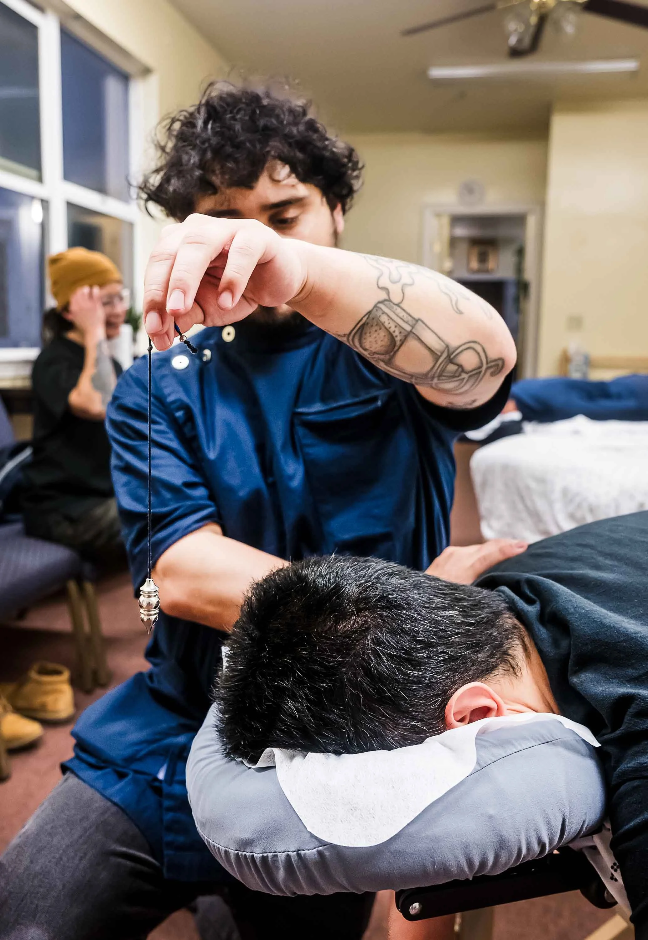 A person receiving a massage or chiropractic adjustment, lying face down on a table, with another person standing beside them and holding a string or cord, in an indoor setting with people and furniture in the background.