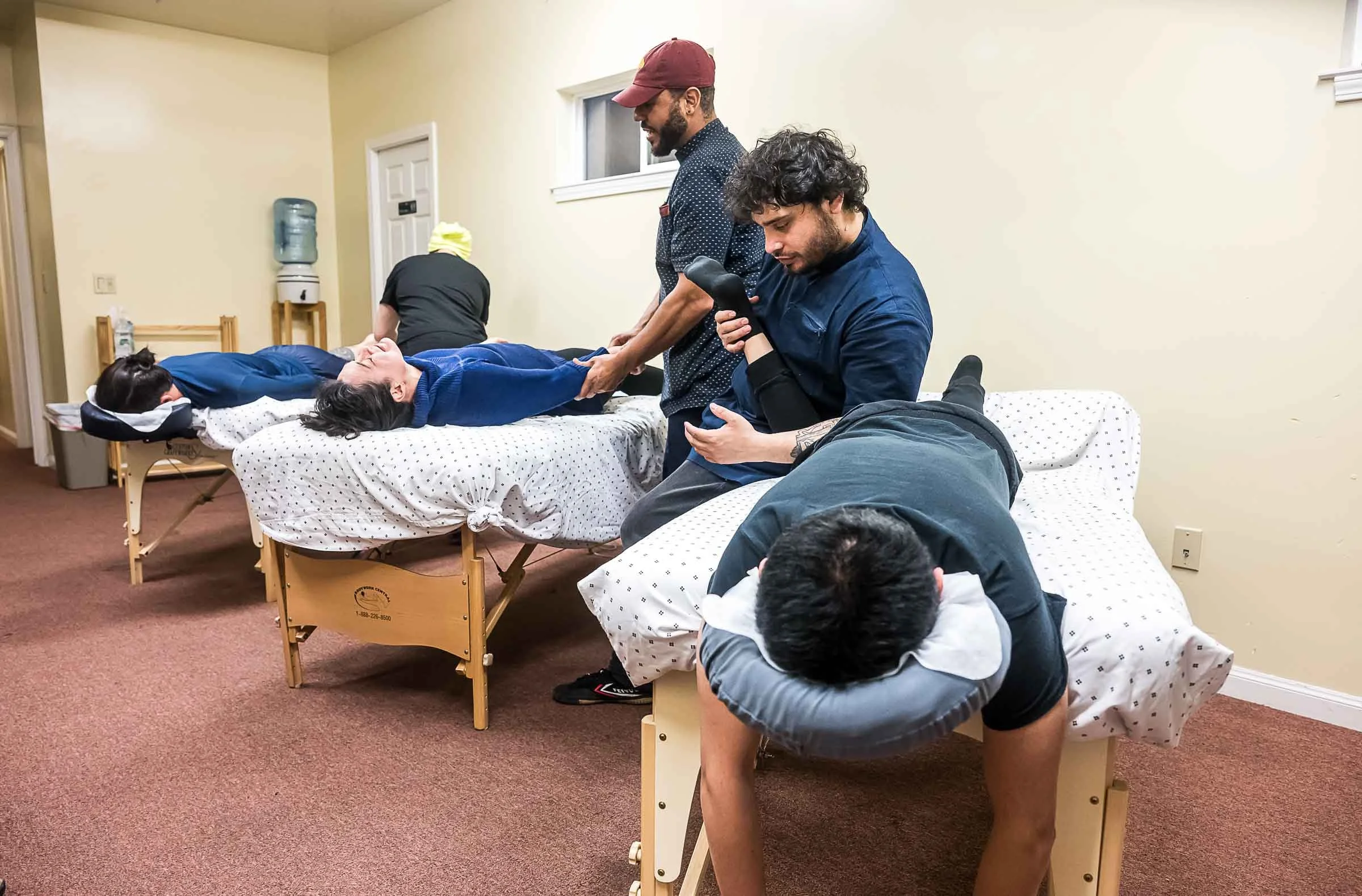 Group of people participating in a massage class, practicing massage techniques on massage tables, with a room featuring beige walls and carpeted floor.