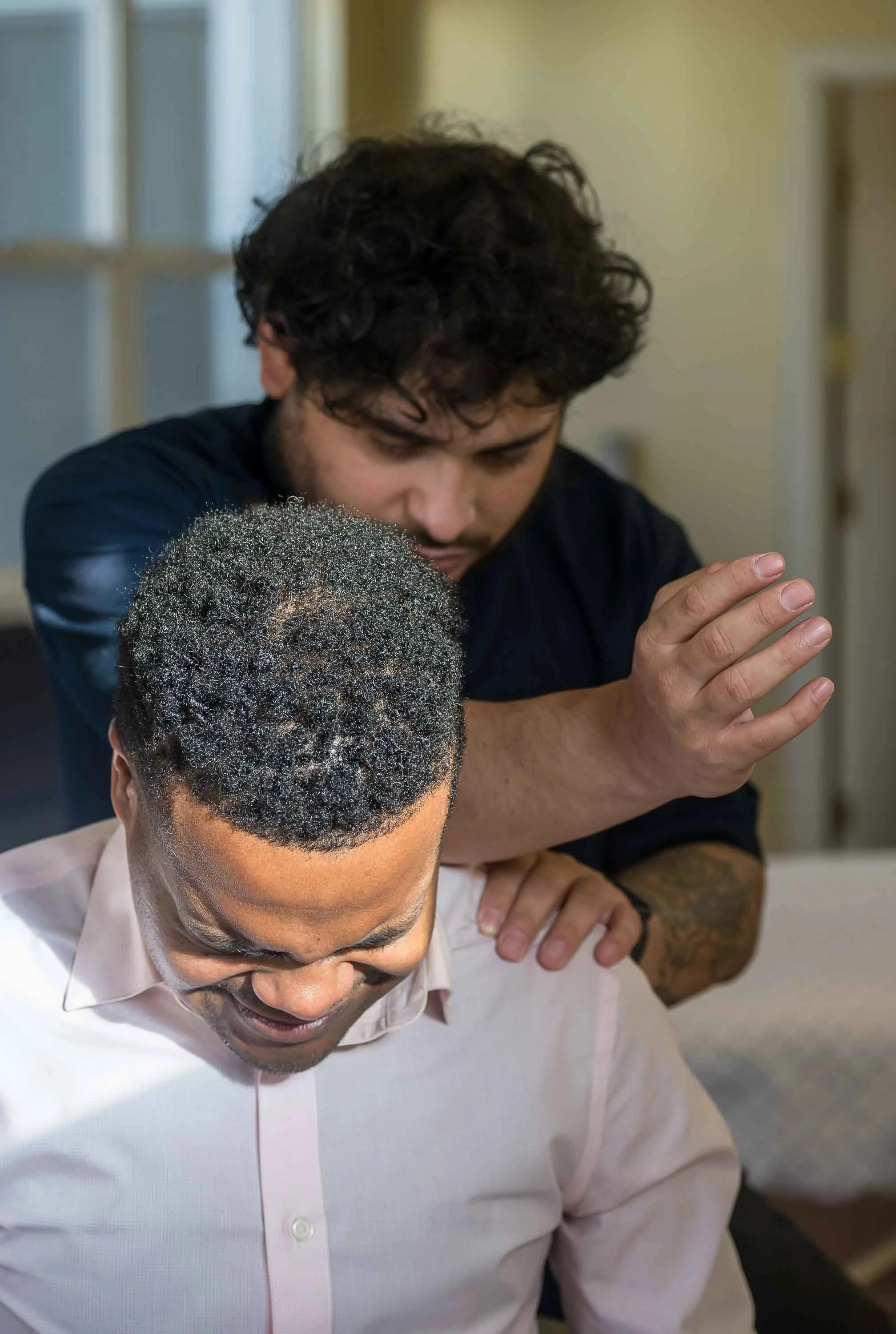 Two men, one with dark, curly hair and the other with short, graying hair and glasses, are in a close interaction. The man with glasses is sitting down, and the other man is standing behind him, with his hands clasped in a prayer-like gesture, possibly praying or showing respect.