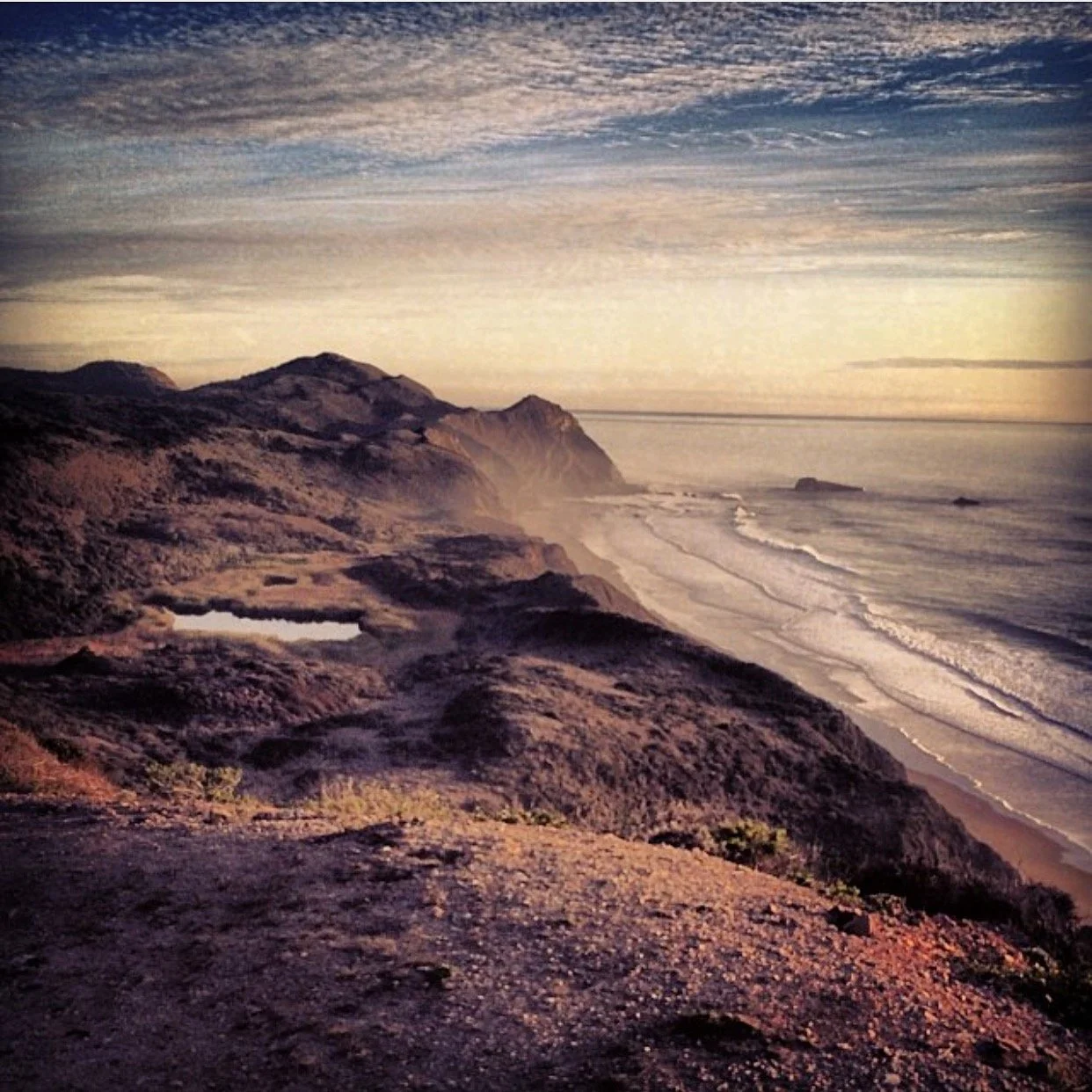 Coastal landscape with rugged cliffs, sandy beach, ocean waves, and cloudy sky at sunset or sunrise.