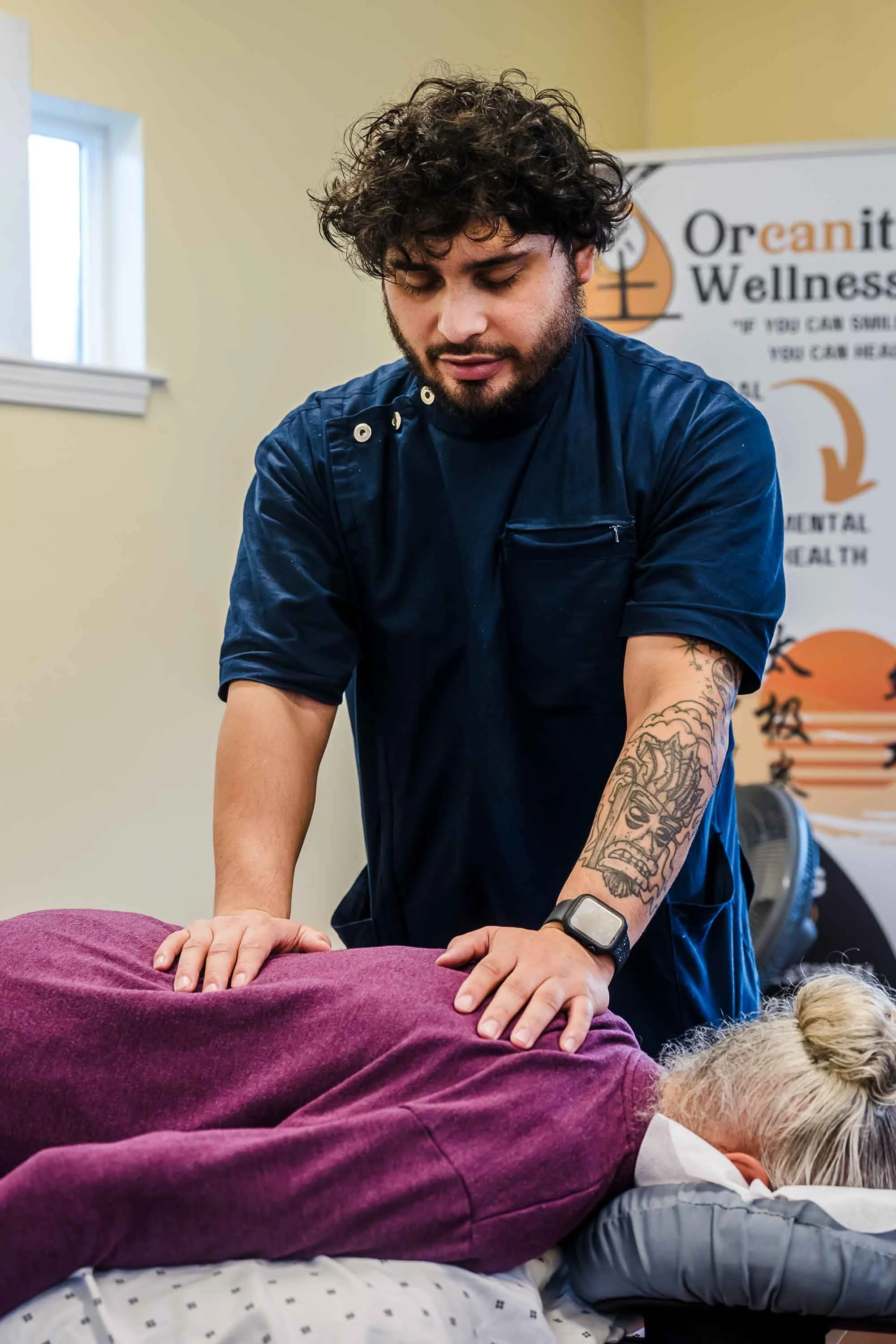A man with curly hair and tattoos performs chiropractic adjustment on a patient lying face down, dressed in a maroon shirt, in a wellness clinic with a banner in the background.