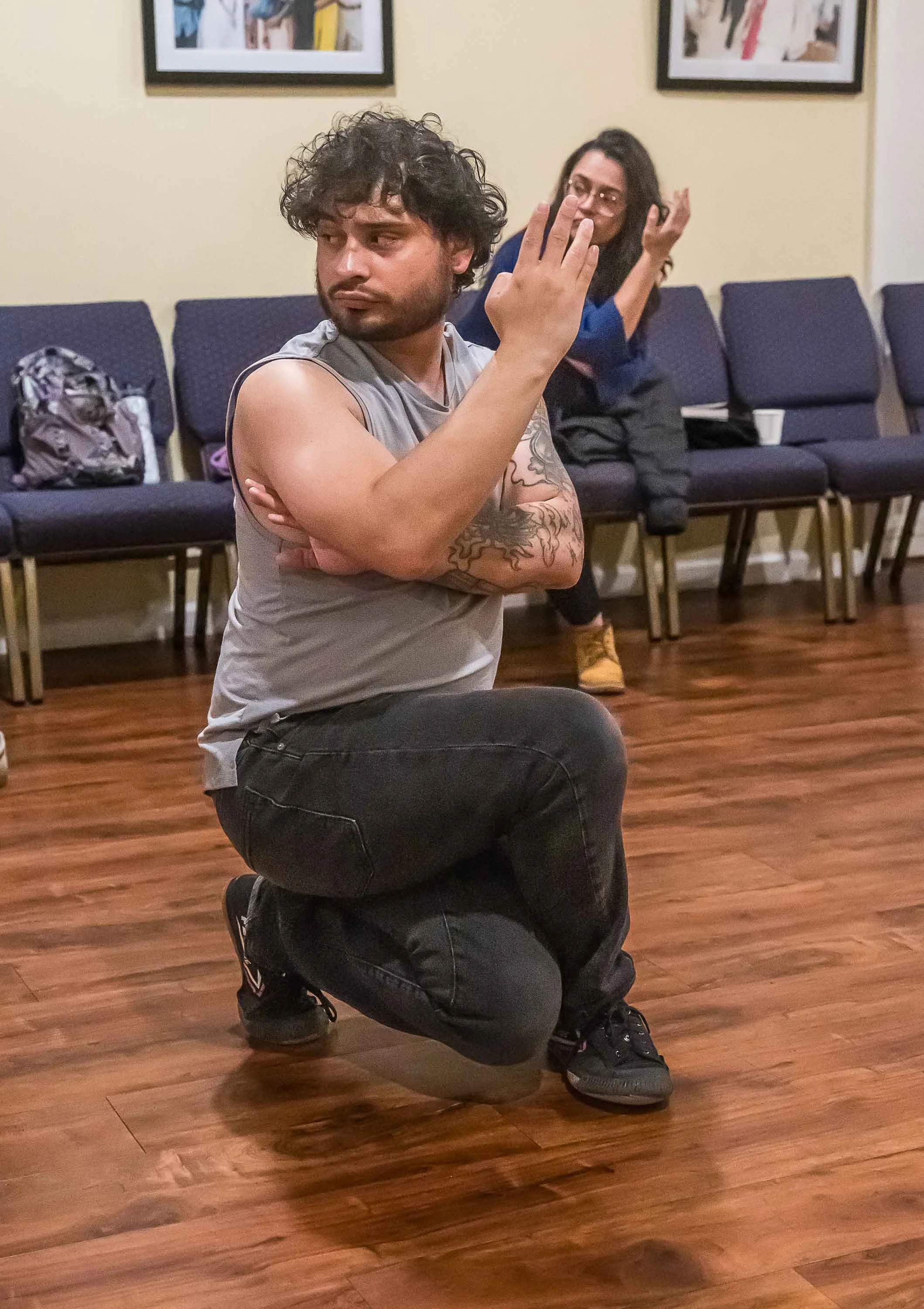 A man kneeling on one knee with his arm crossed and his other arm raised, making a gesture with his hand. A woman sitting on a chair behind him, with glasses and long dark hair, appears to be talking or explaining something. The setting looks like a community center or classroom with chairs, framed pictures on the wall, and a wooden floor.