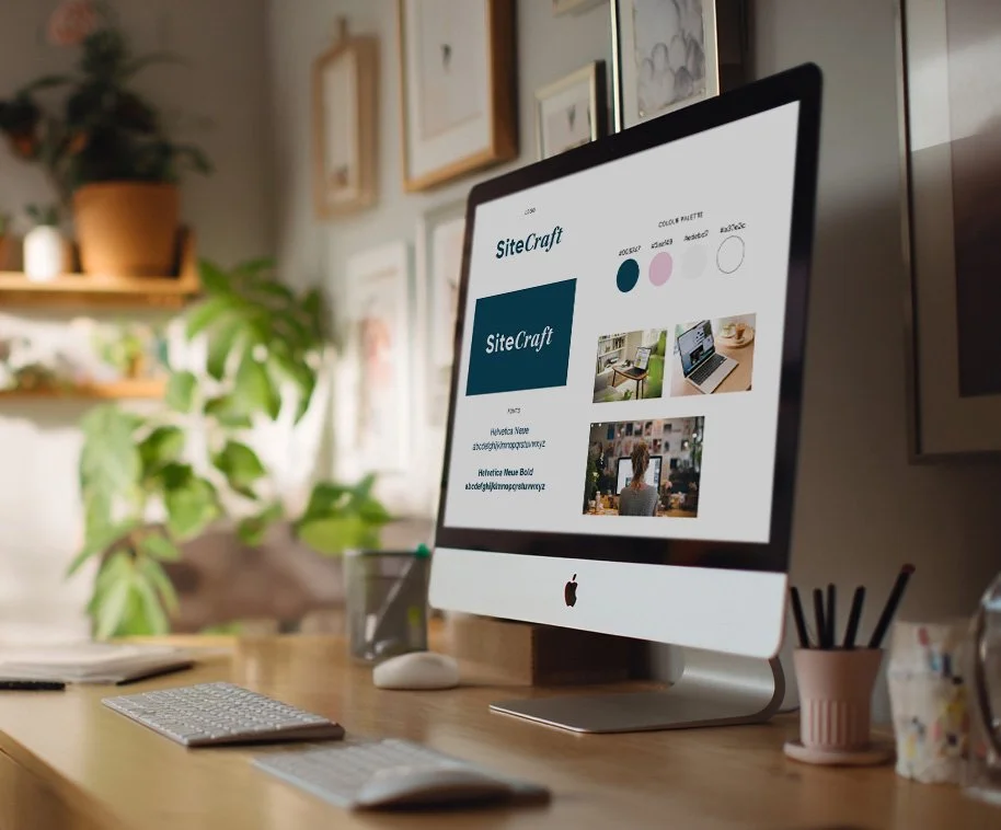 An Apple iMac computer on a wooden desk displaying a website called SiteCraft, with a keyboard, mouse, pencils in a cup, and a notebook nearby, in a room with framed artwork and plants.