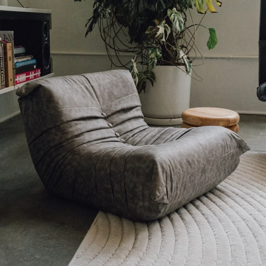 A cozy, gray, velvety floor chair on a beige rug in a living room with a potted plant, a small wooden stool, a bookshelf, and a speaker.