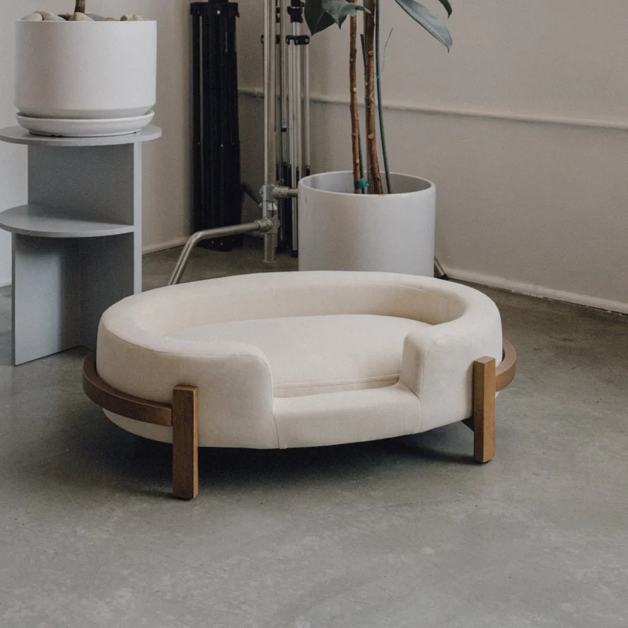 A modern, oval-shaped cream-colored pet bed with wooden legs, placed on a concrete floor. In the background, there is a round white side table with a potted plant, a large white planter with a leafy plant, and some metal pipes and equipment.