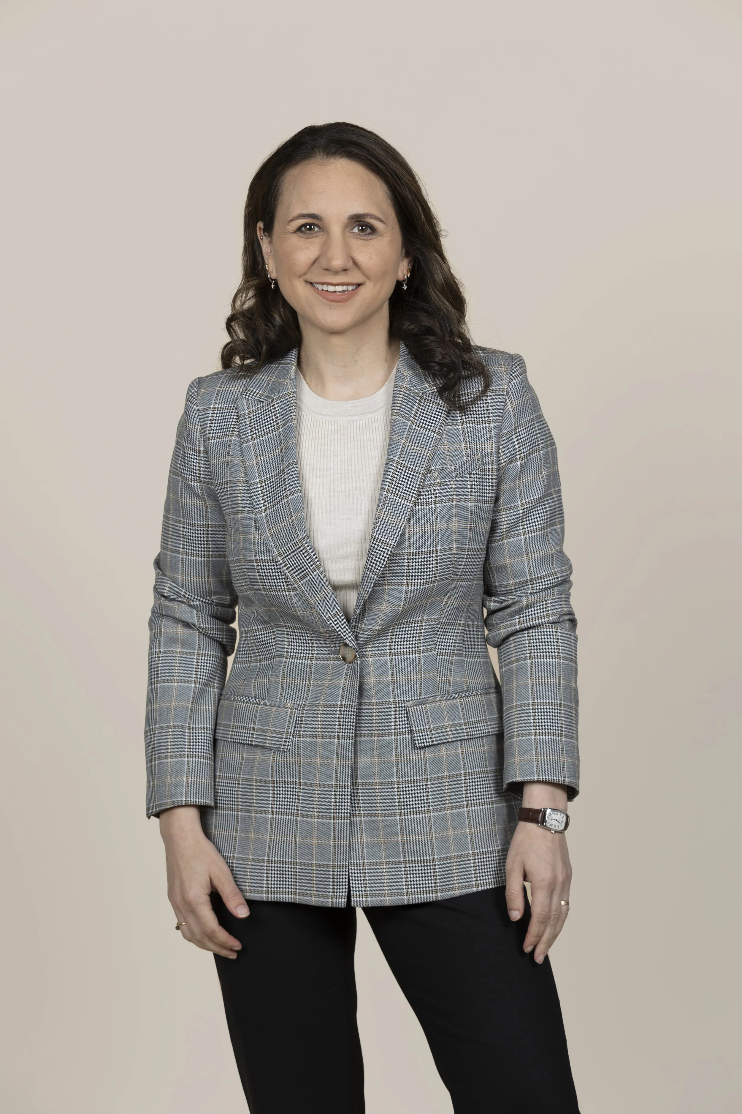 Woman with dark brown hair wearing a plaid blazer and black pants standing against a beige background.