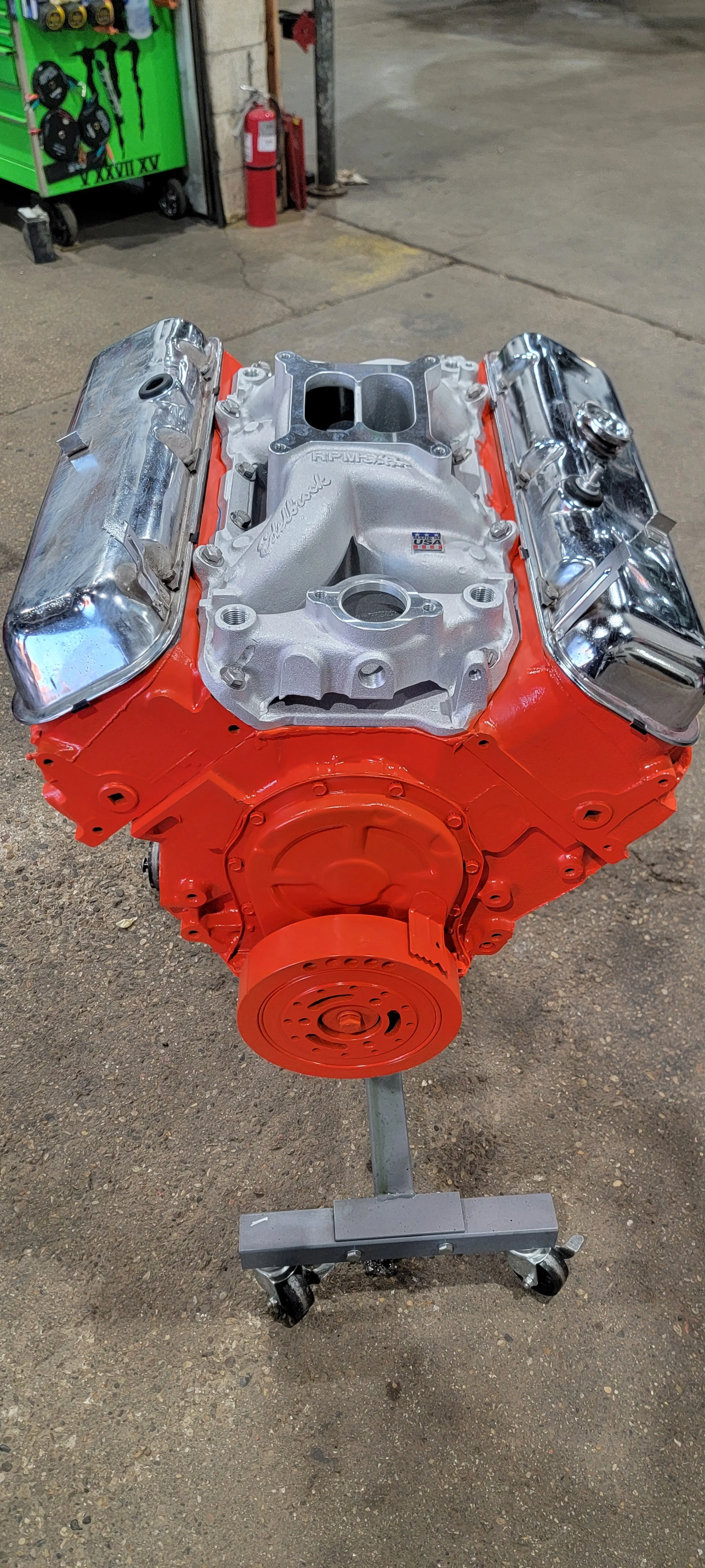 A large, orange V8 engine on a wheeled stand inside a workshop or garage.