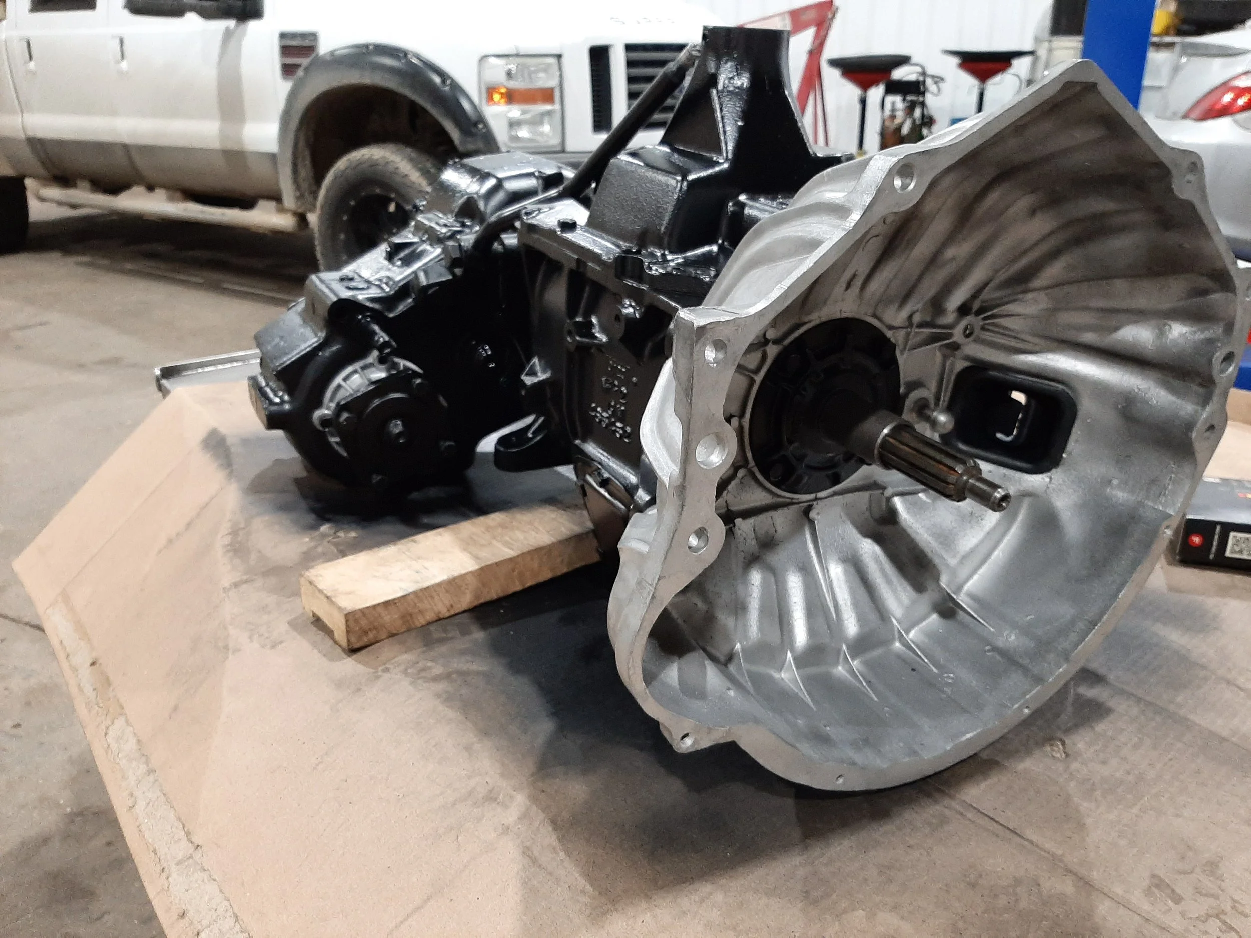 An automobile transmission is placed on a wooden block on the floor of a garage, with a truck and various tools visible in the background.