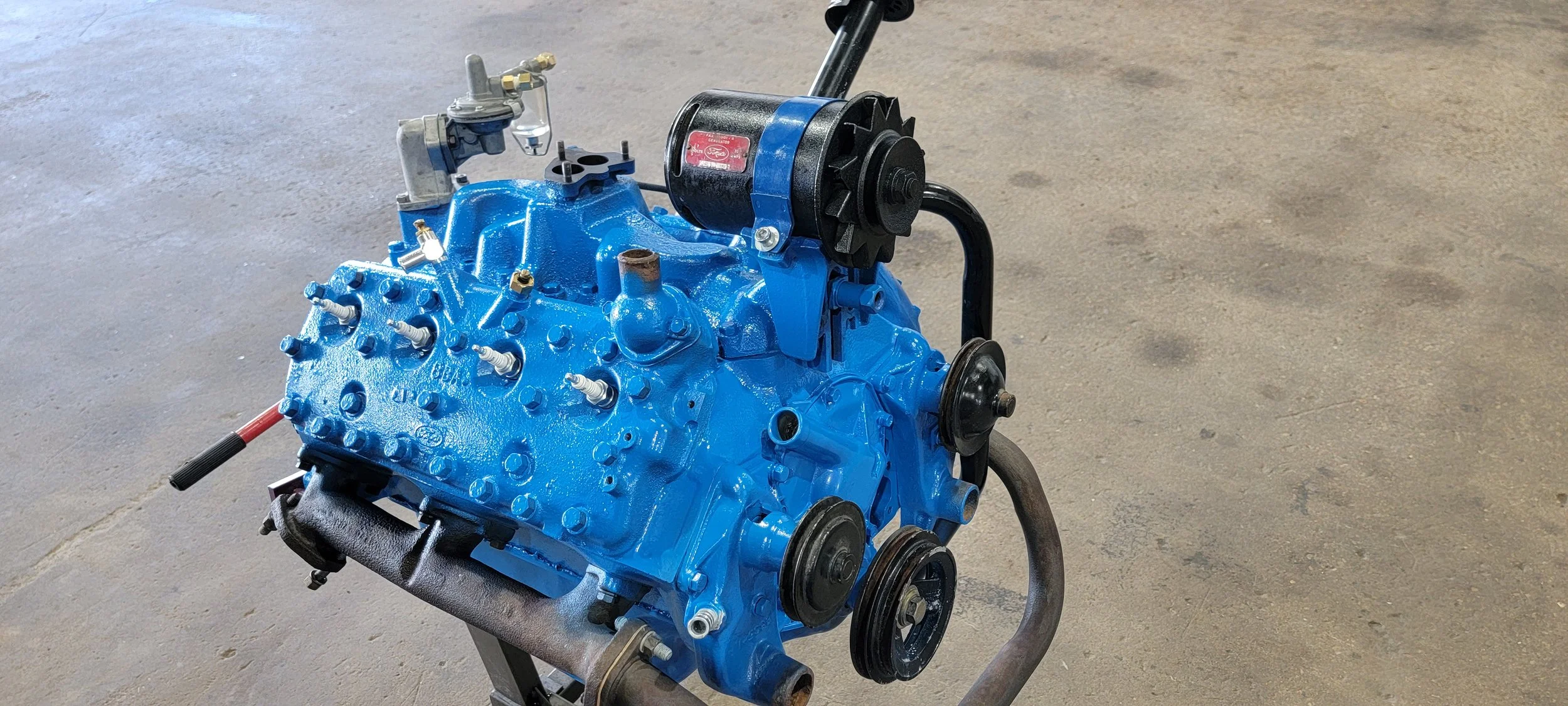 An engine block painted blue with black pulleys and a black motor attached, mounted on a stand against a concrete floor.