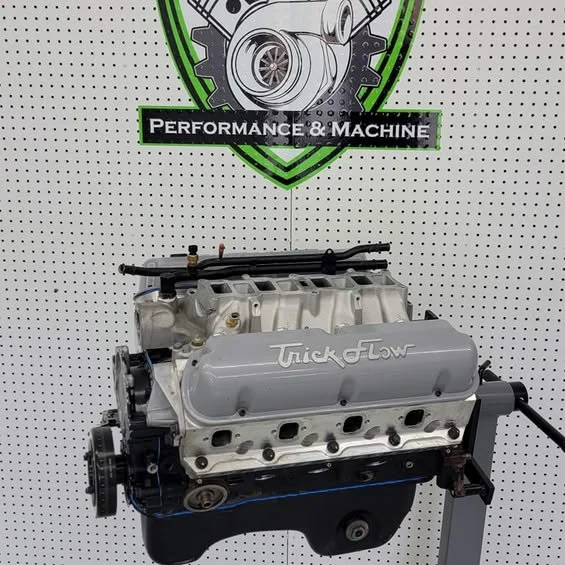 A car engine mounted on a stand against a white pegboard background with a performance and machine logo. The engine has a silver valve cover with 'Trick Flow' written on it.
