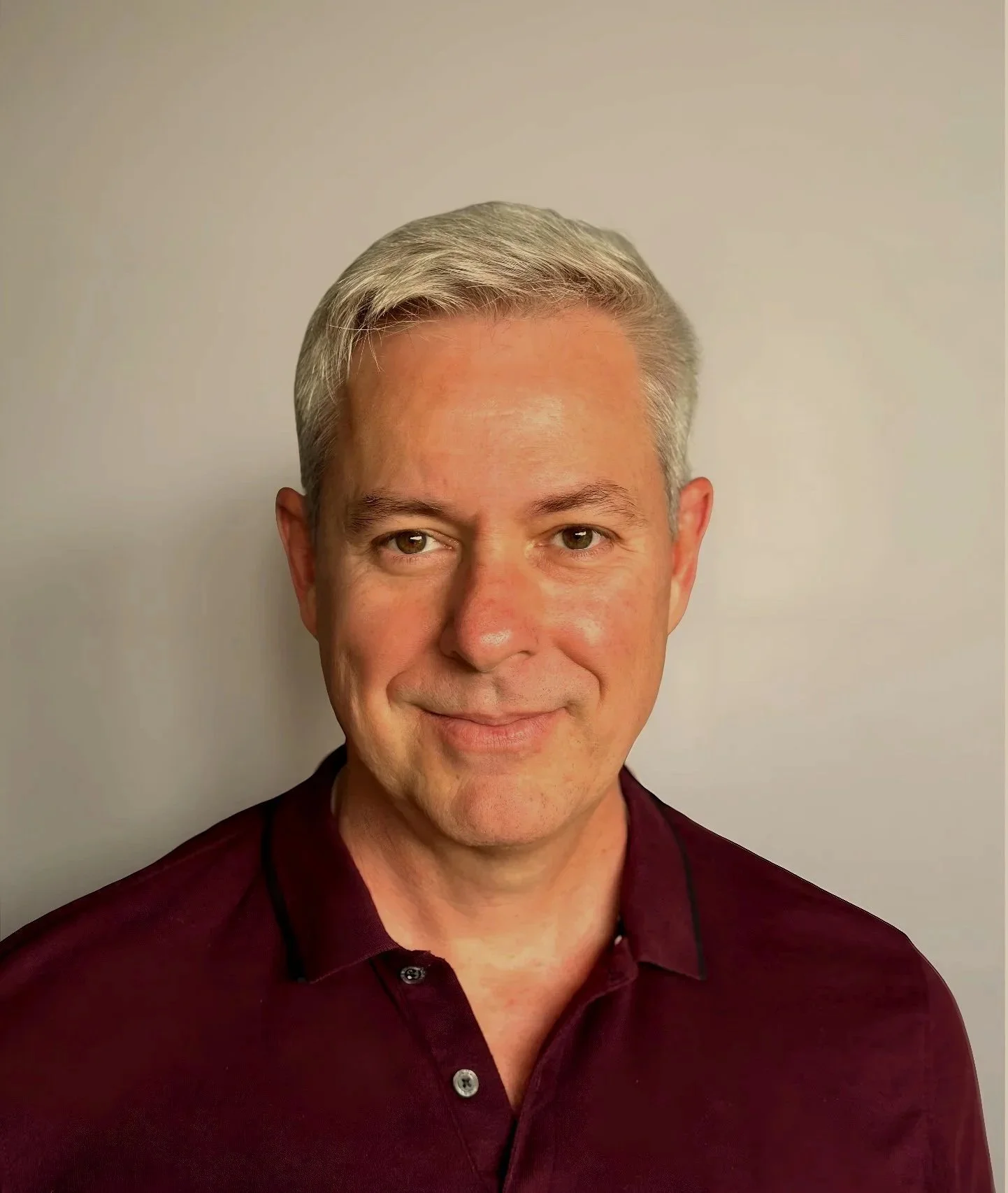 A man with grey hair wearing a maroon collared shirt, standing against a plain light-colored wall, smiling slightly.
