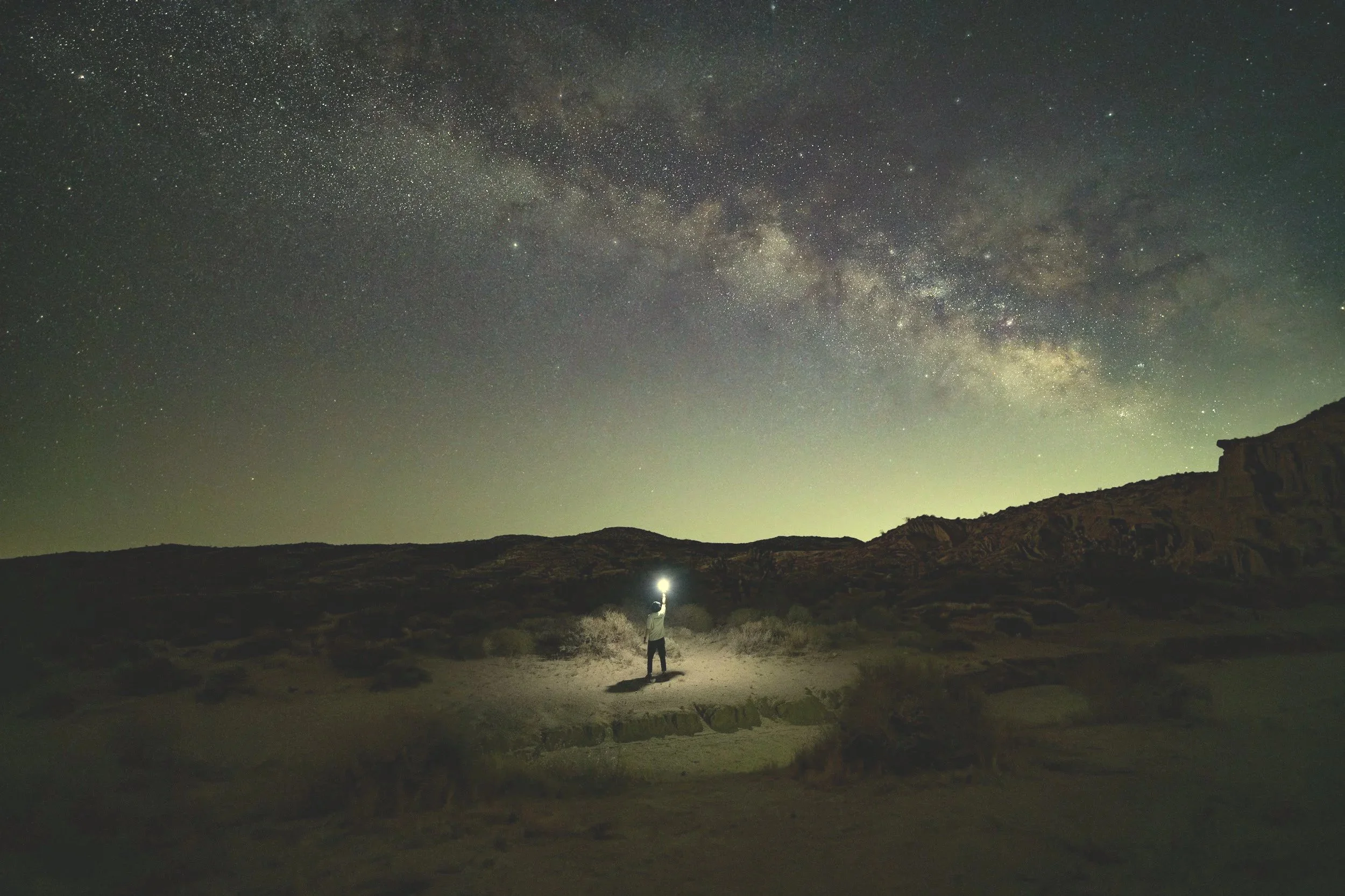 A person standing in a desert landscape at night, holding a flashlight with the Milky Way galaxy visible above in the sky.