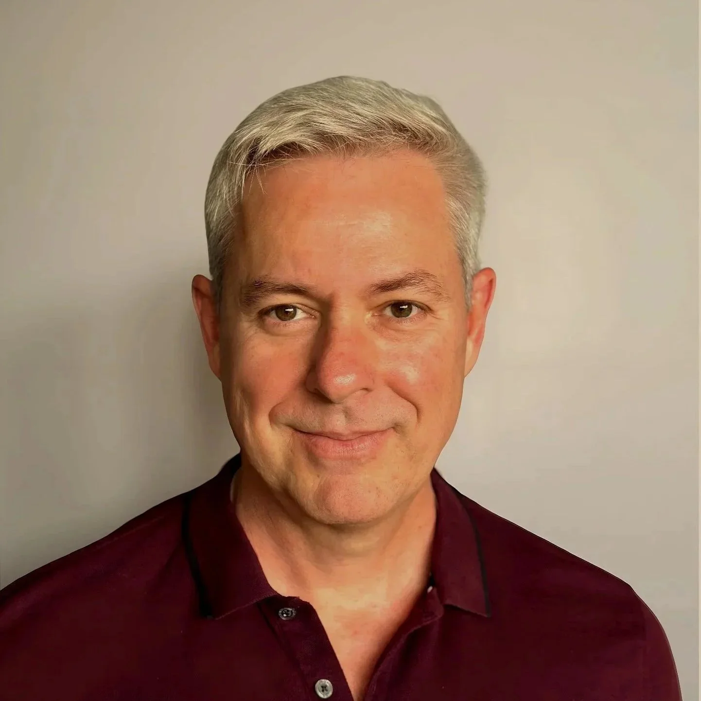 A middle-aged man with gray hair wearing a maroon collared shirt, smiling softly against a plain light-colored background.