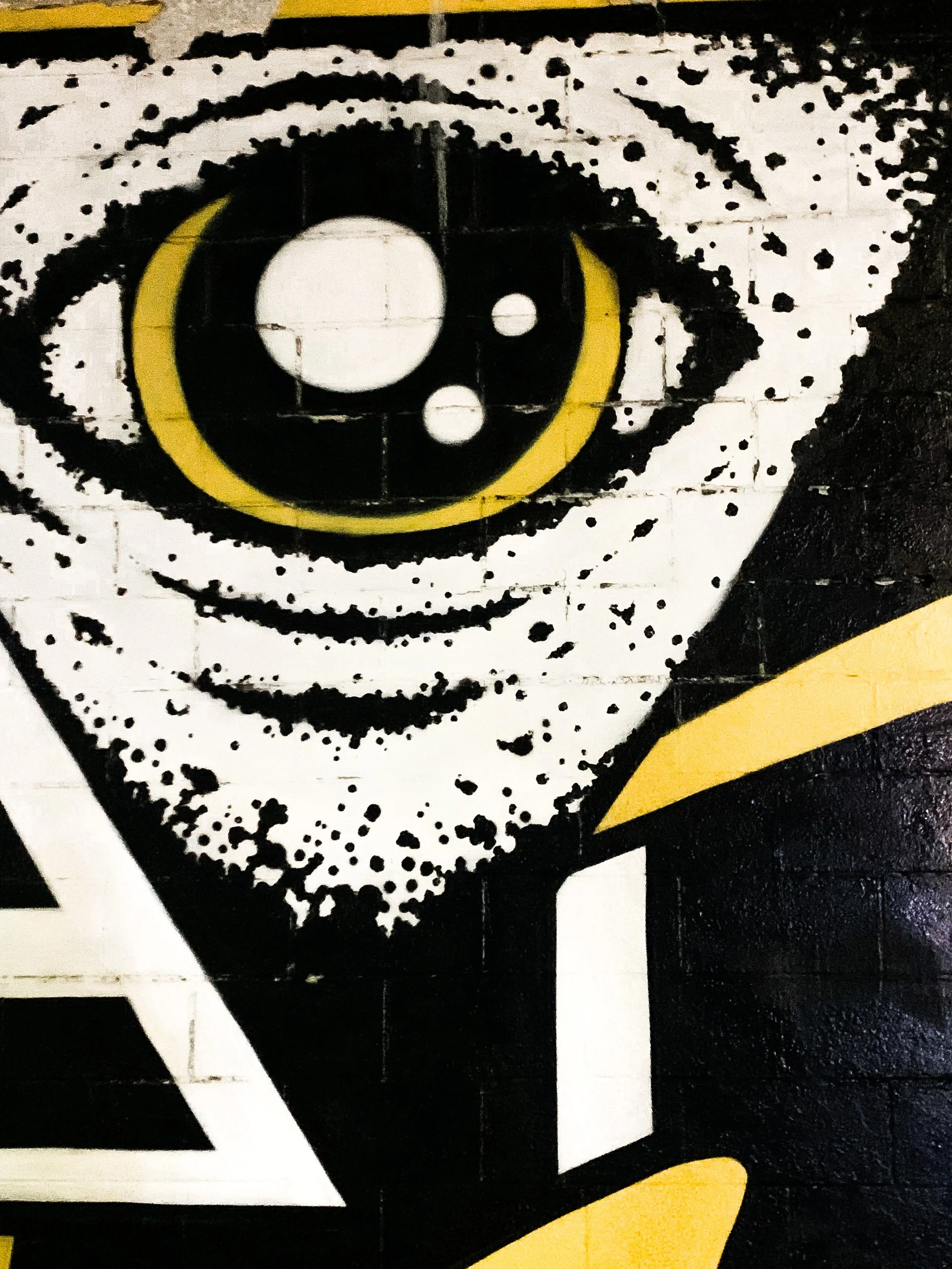 A close-up of a graffiti mural featuring a large eye with a yellow iris and black pupil, decorated with white dots and a white circle, set against a black and white background with geometric shapes.
