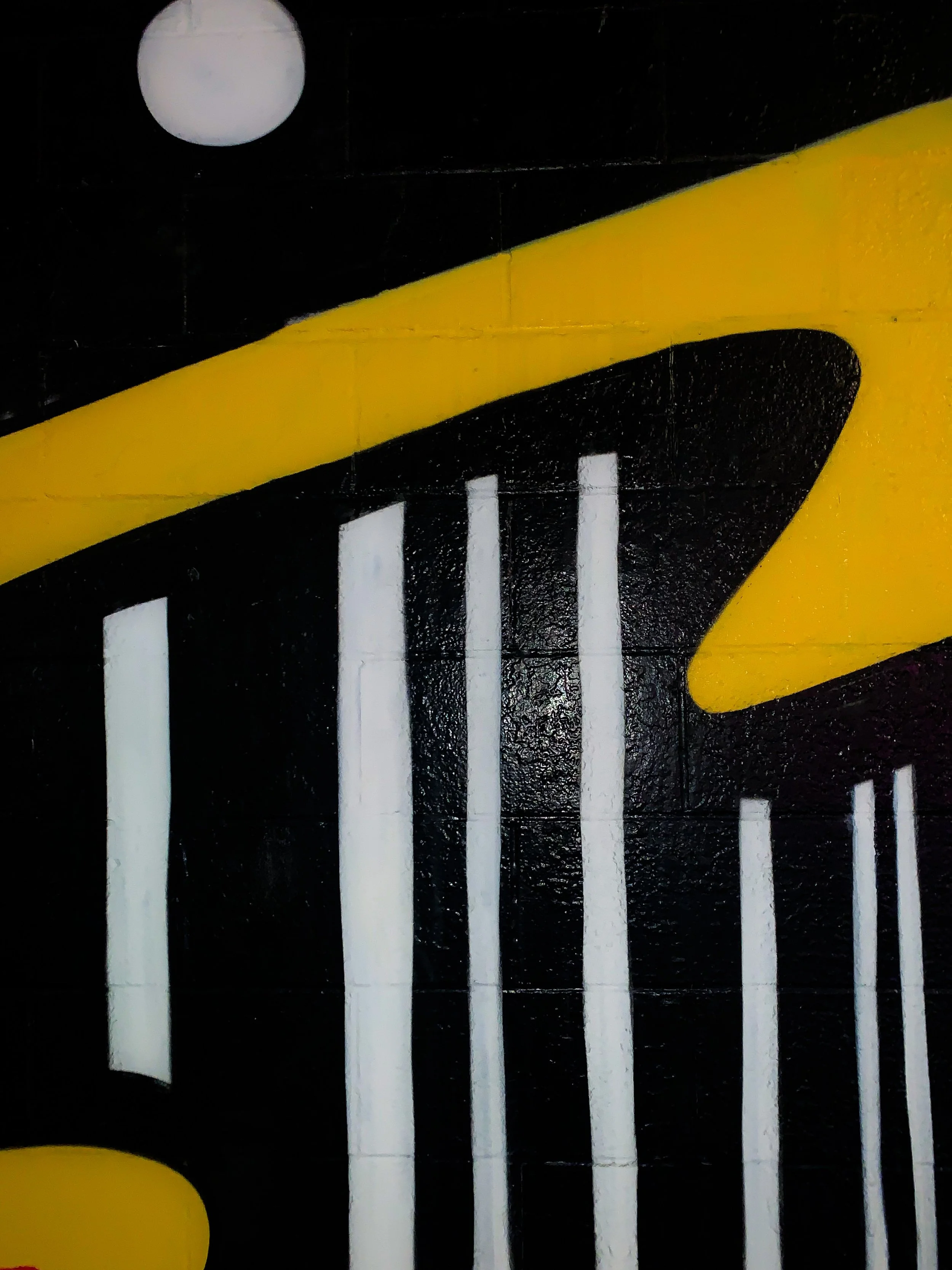 Close-up of graffiti art with white vertical lines, yellow and black abstract shapes, on a black brick wall, with a white circular object in the upper left corner.
