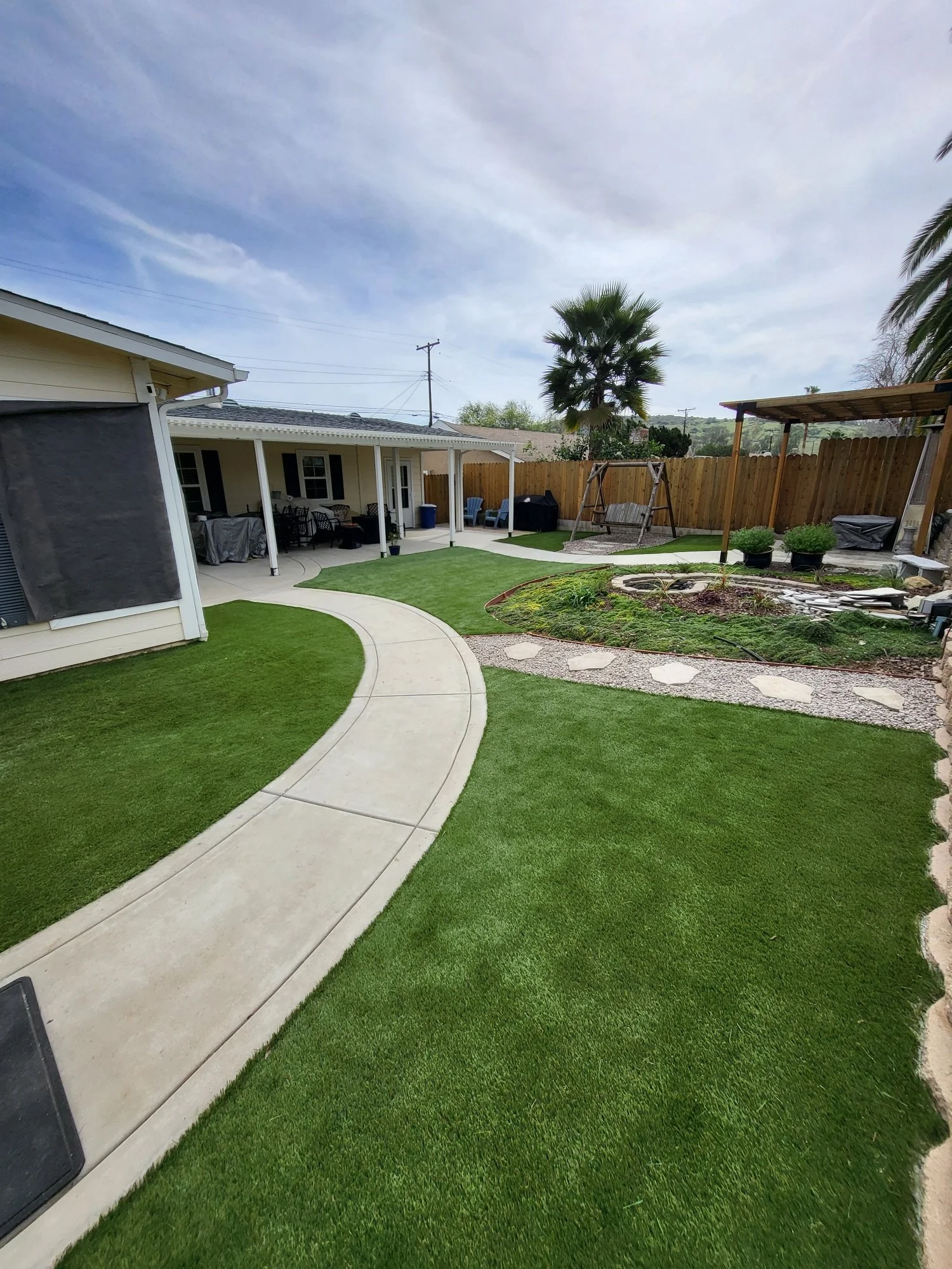Backyard with a curved concrete pathway, lush green grass, a covered patio with outdoor furniture, a swing, potted bushes, and a wooden fence, under a partly cloudy sky.