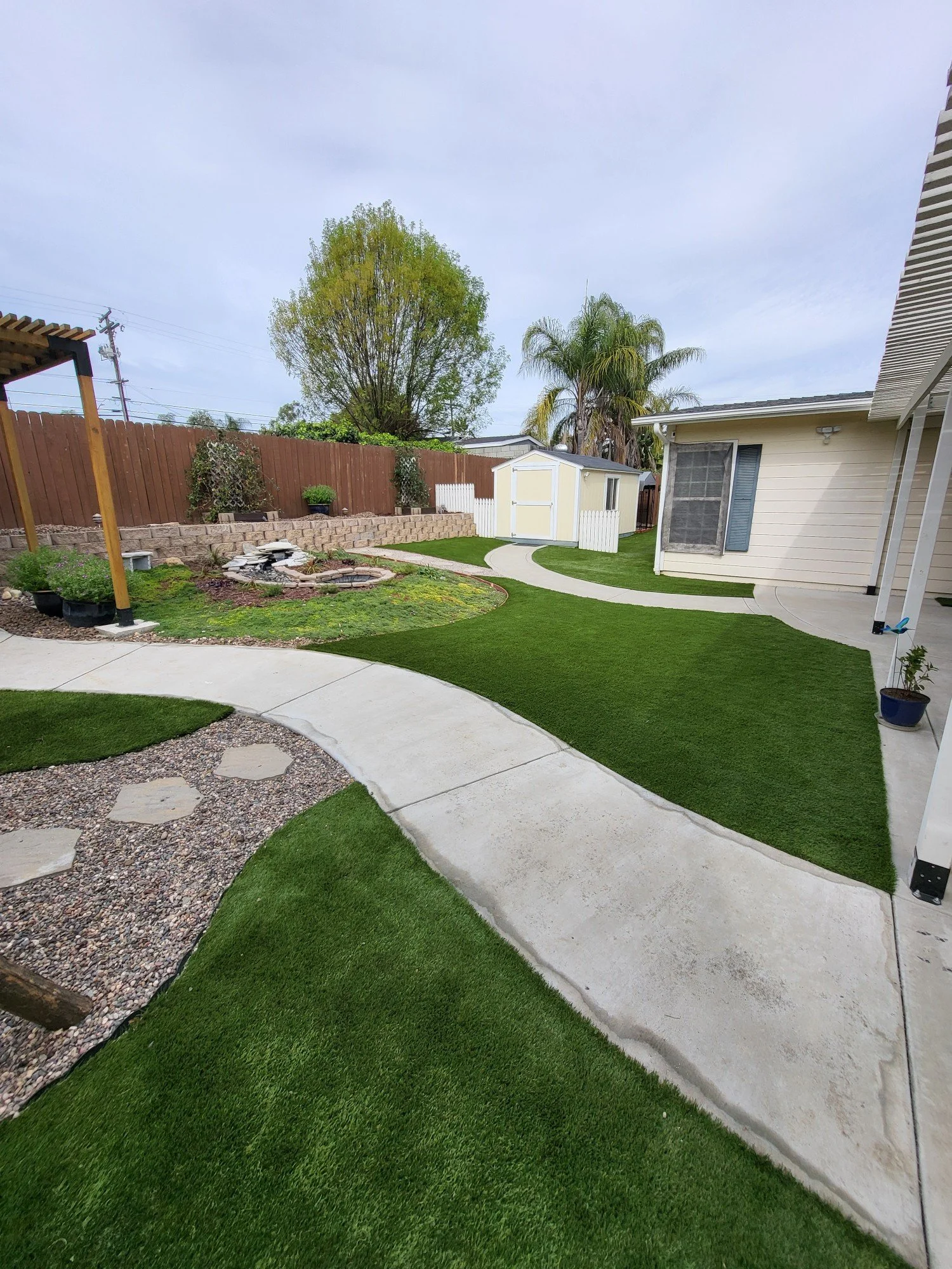 A backyard with curved concrete walkways, lush green grass, a small raised garden bed with plants, a white shed with a yellow door, trees, and a wooden fence.