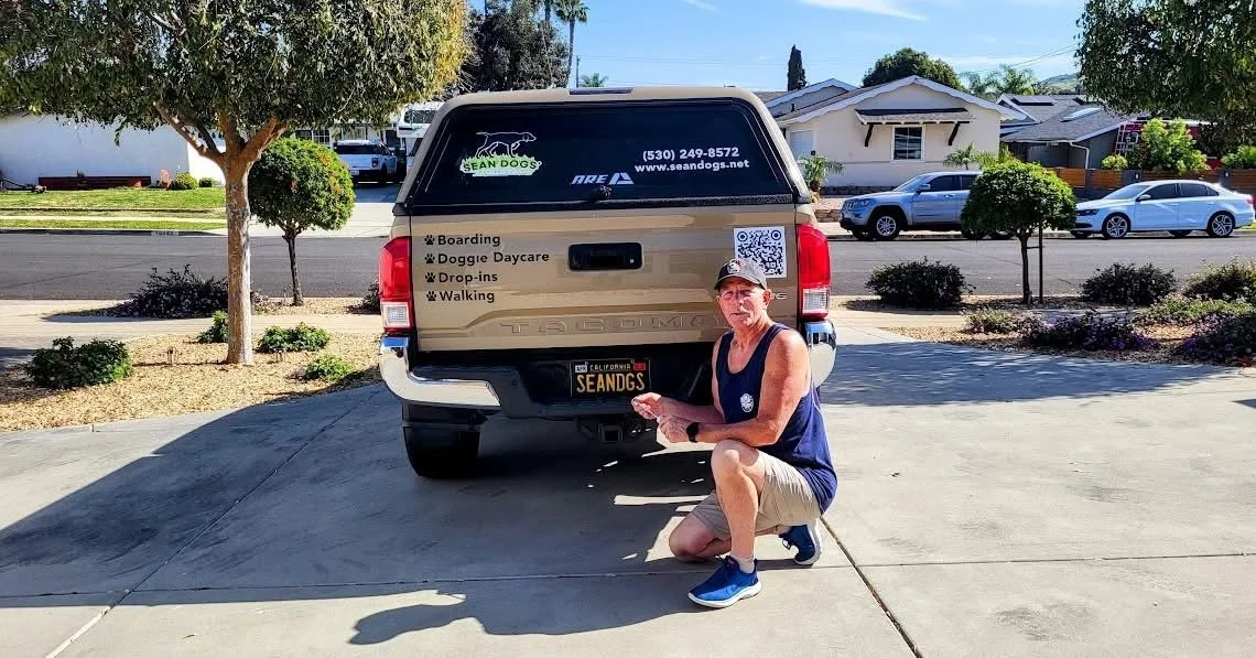 A man kneeling in front of a dog transport truck parked in a driveway in a suburban neighborhood. The truck has signage for SEANDOGS, a pet service company, including a website and phone number, and lists services like boarding, doggie daycare, drop-ins, and walking.