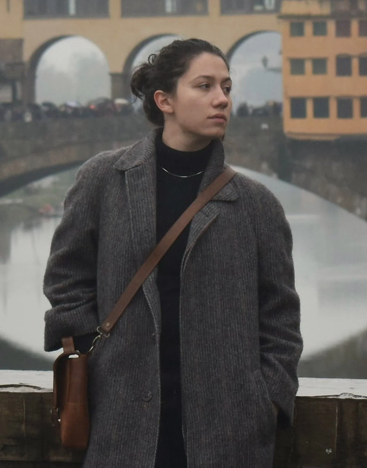 Person standing in front of Ponte Vecchio, Florence, wearing a gray coat and carrying a brown bag.