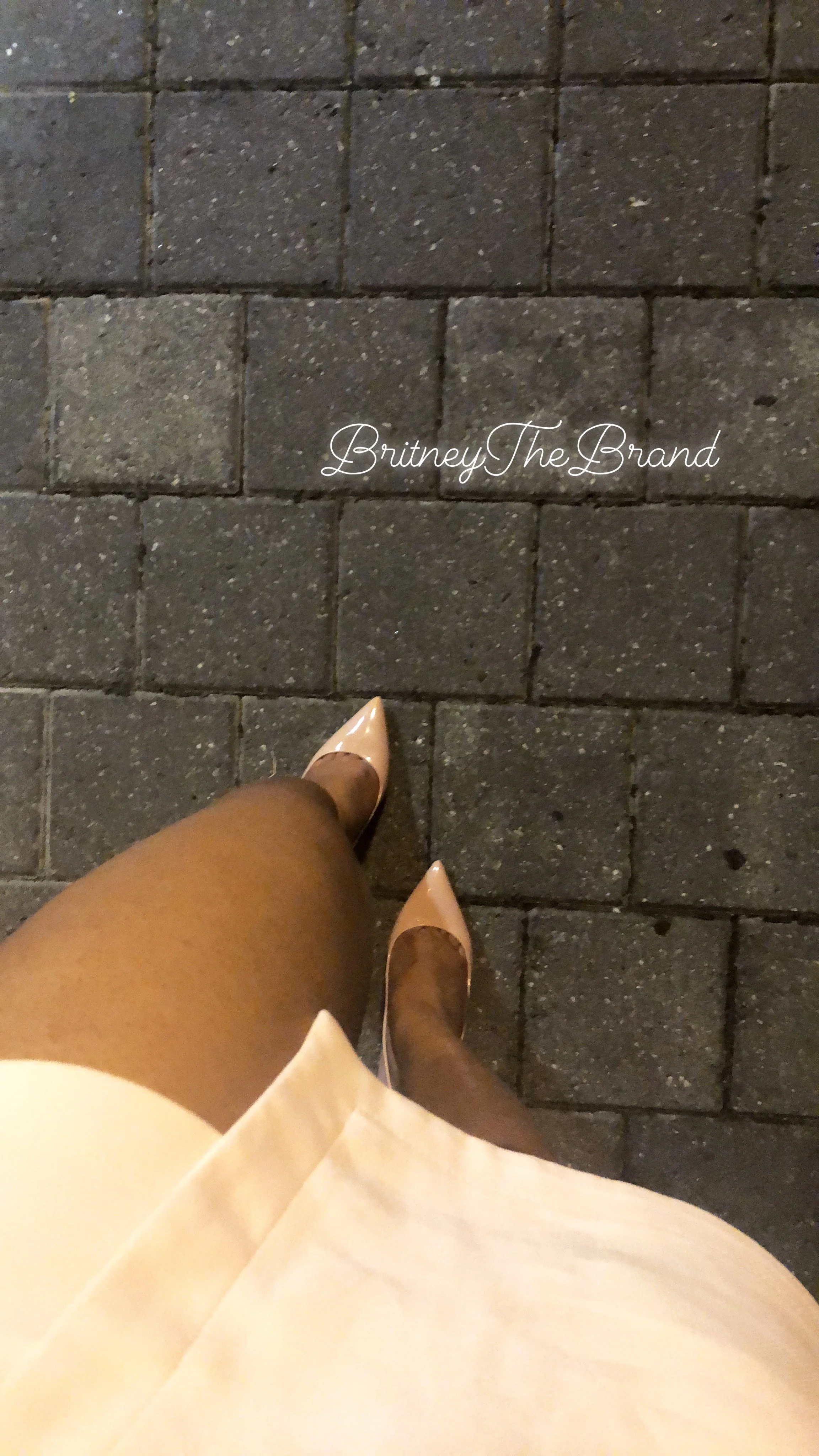Photo showing a view of a person's legs wearing nude-colored high heels and a beige dress, walking on a brick sidewalk at night. The text 'BritneyTheBrand' is overlaid on the image.