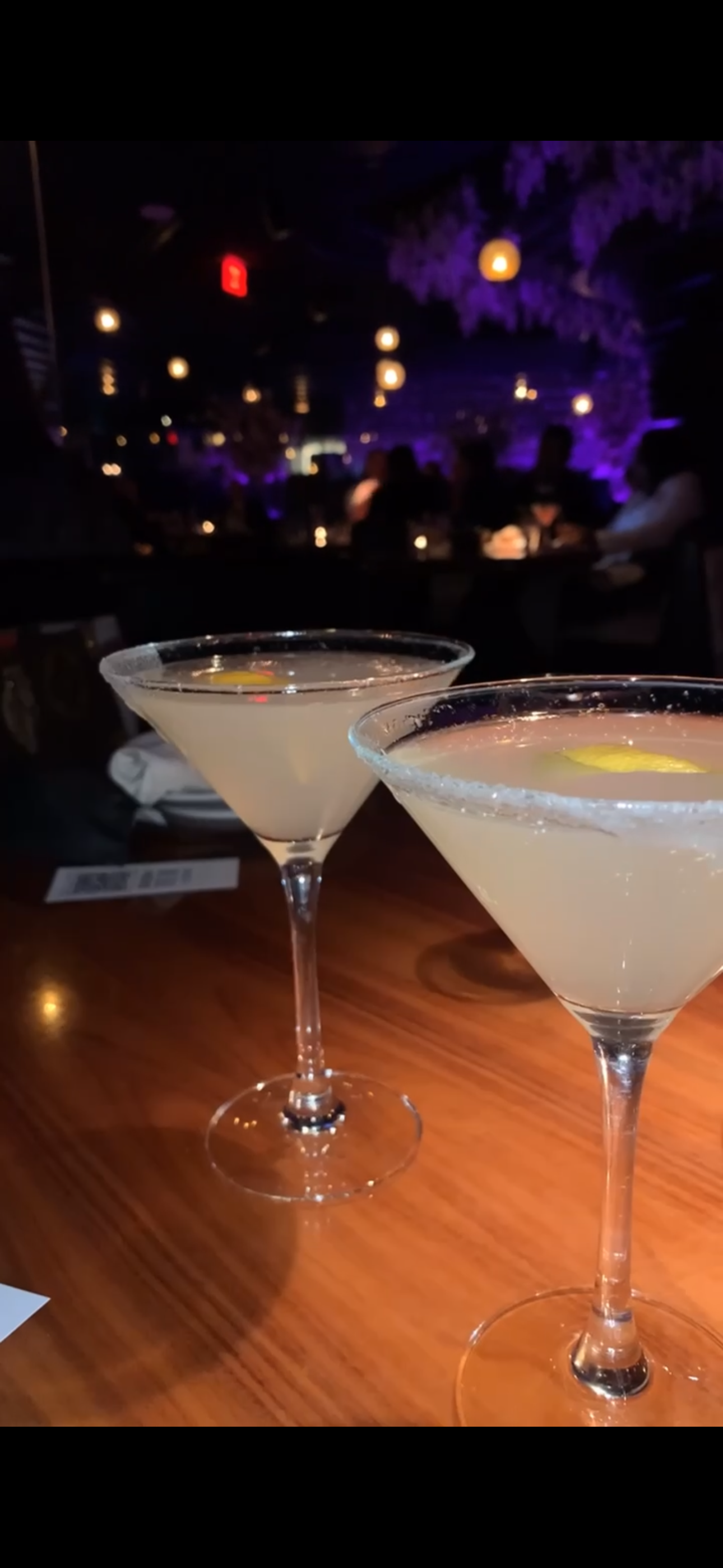 Two cocktails in martini glasses on a wooden table in a dimly lit bar or nightclub.
