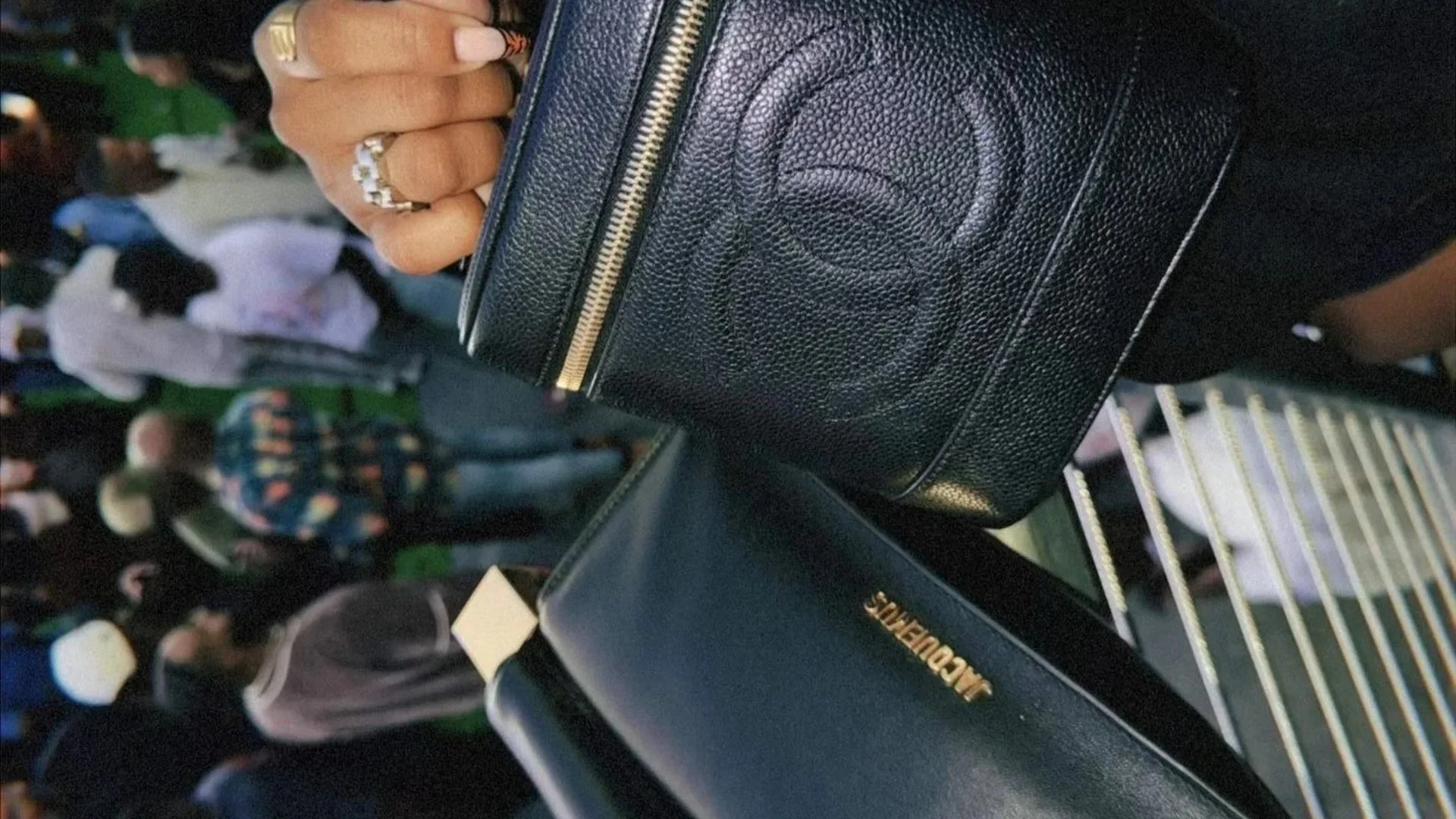 Close-up of a hand holding a black designer handbag with gold zipper details, set against a crowded background of people.