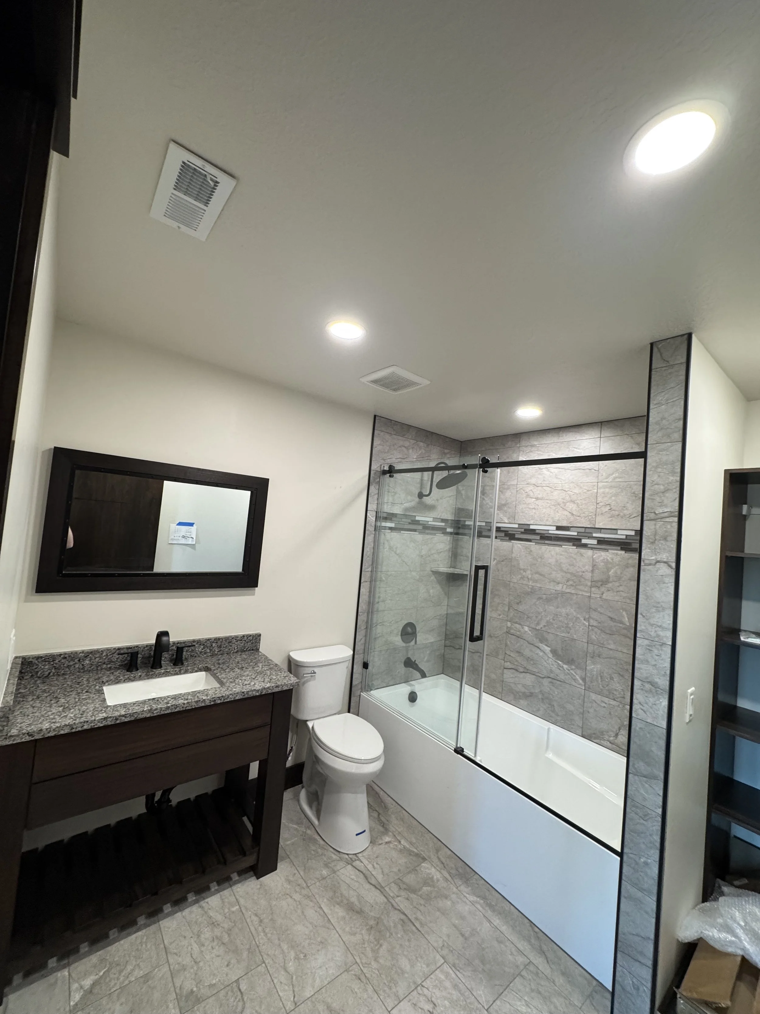 A modern bathroom with a shower-tub combo, toilet, dark wood vanity with granite countertop, mirror, and neutral-colored tiles.