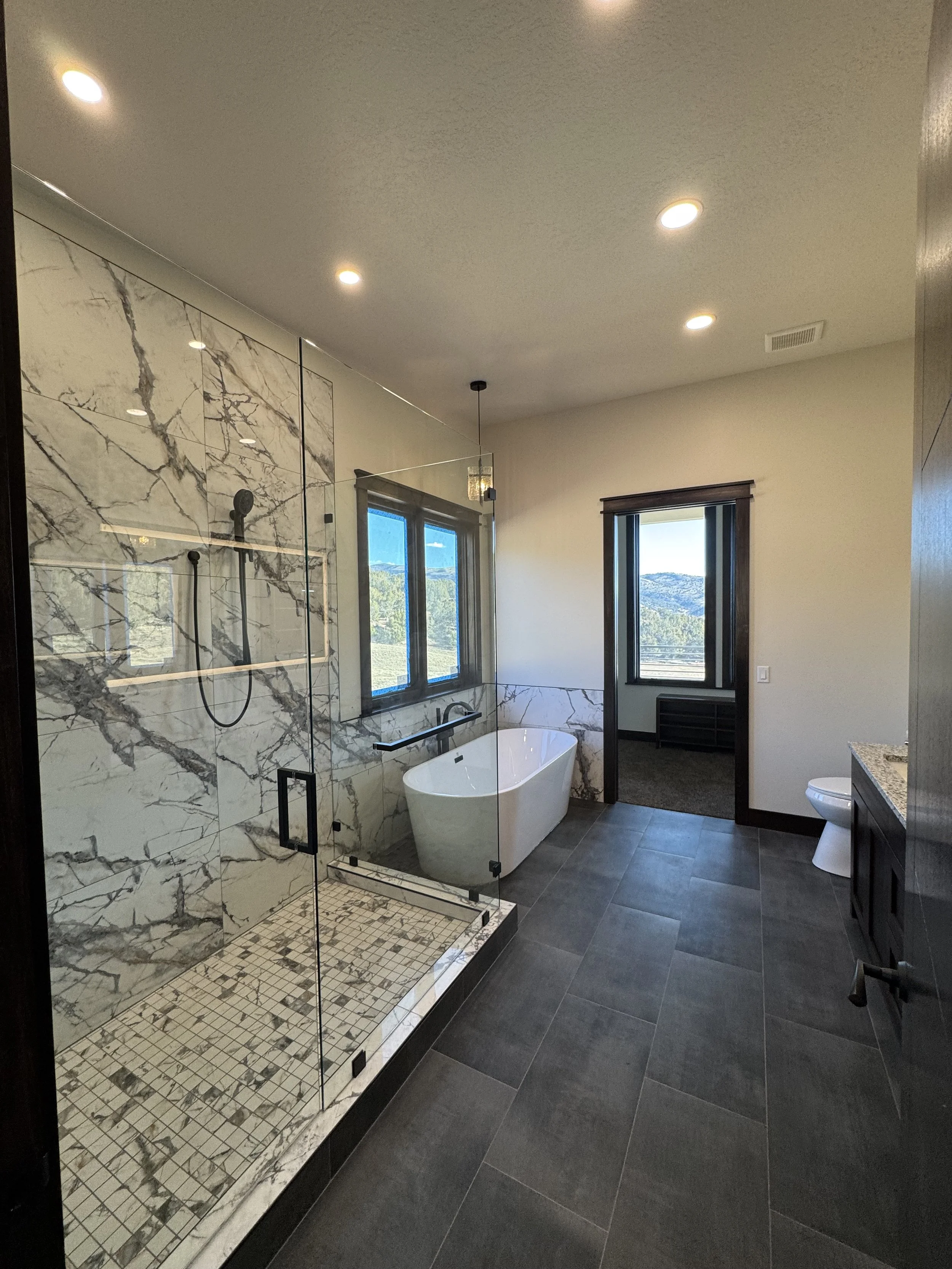 Modern bathroom with a glass-enclosed shower, a freestanding bathtub, two windows, dark floor tiles, and a separate toilet area visible through an open doorway.