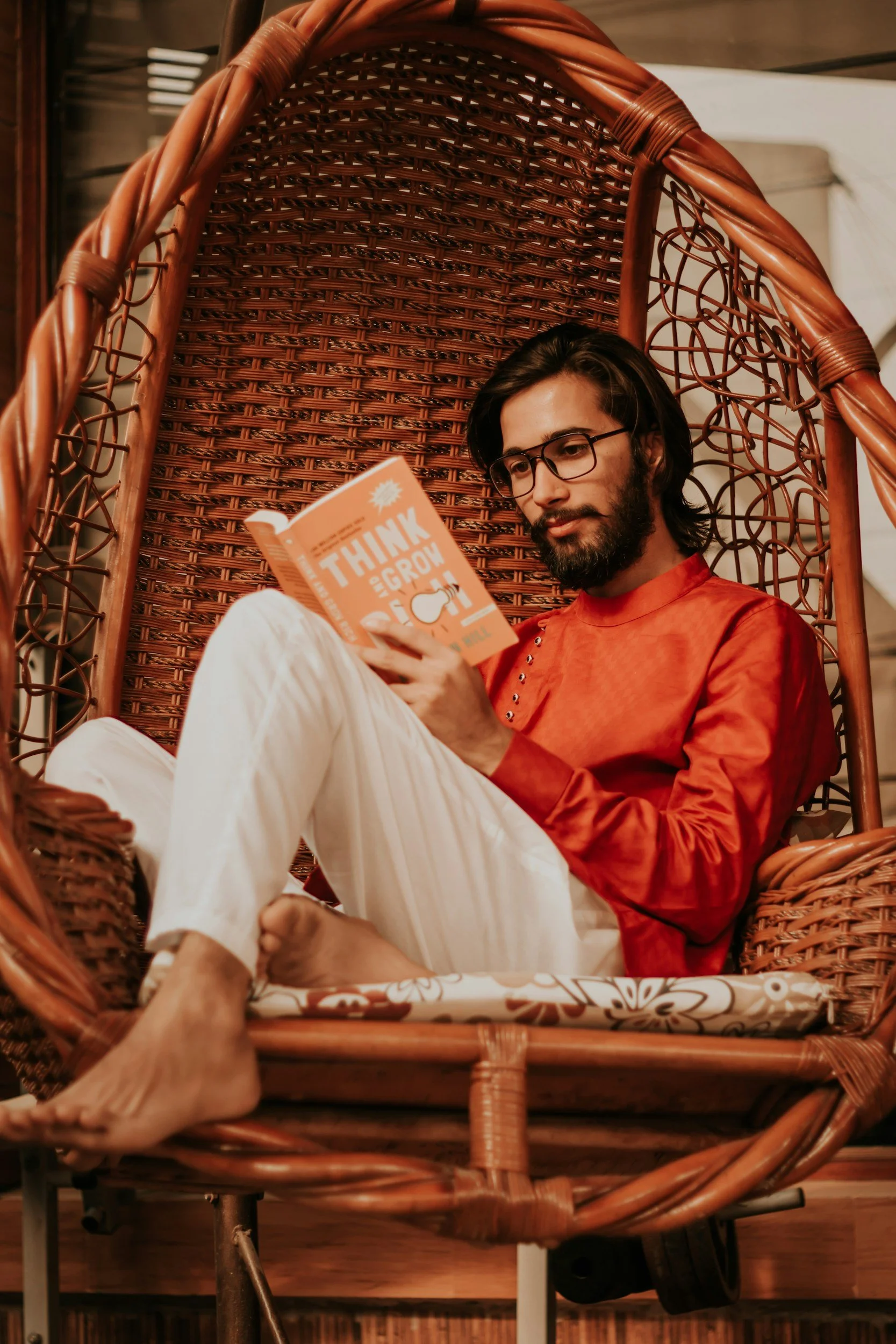 OnlyVibes Open to Solo vibe: A man with long dark hair, glasses, and a beard is sitting in a wicker hanging chair, reading a book titled 'Think and Grow Rich'.