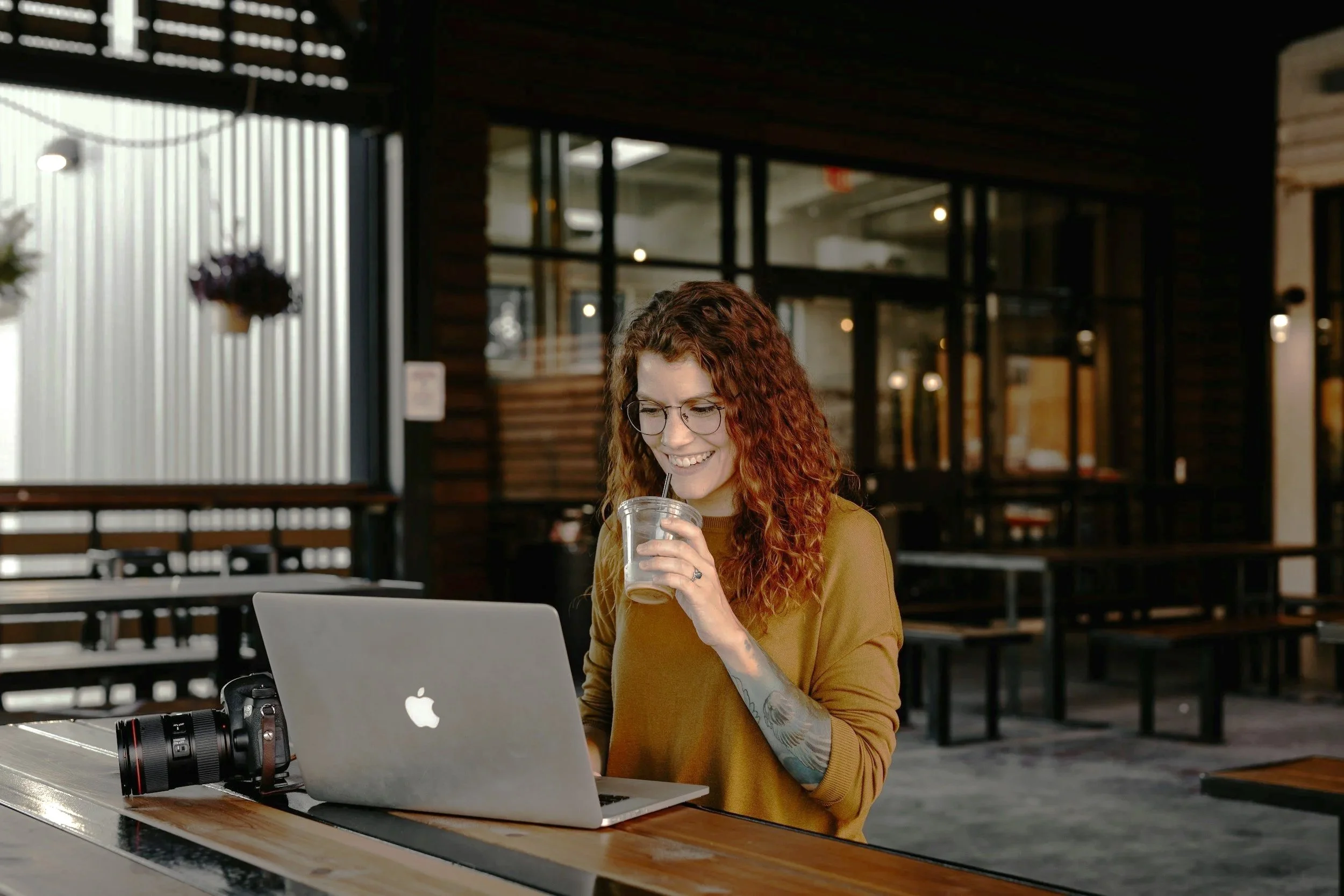 Contact us Page- A woman with curly red hair and glasses sitting at a table in a modern coffee shop, smiling while holding a plastic cup with a straw, in front of a silver MacBook.