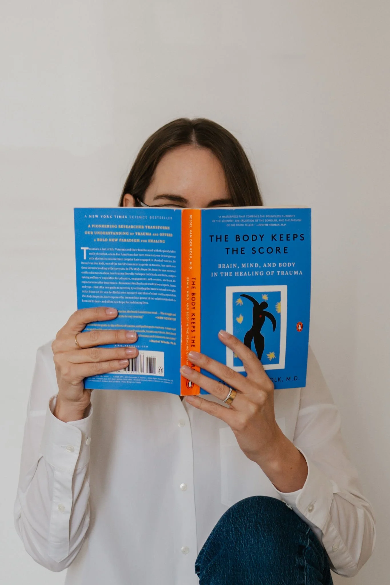 A person wearing a white shirt and blue jeans sitting on a chair, holding a blue book titled 'The Body Keeps The Score' in front of their face, obscuring it.