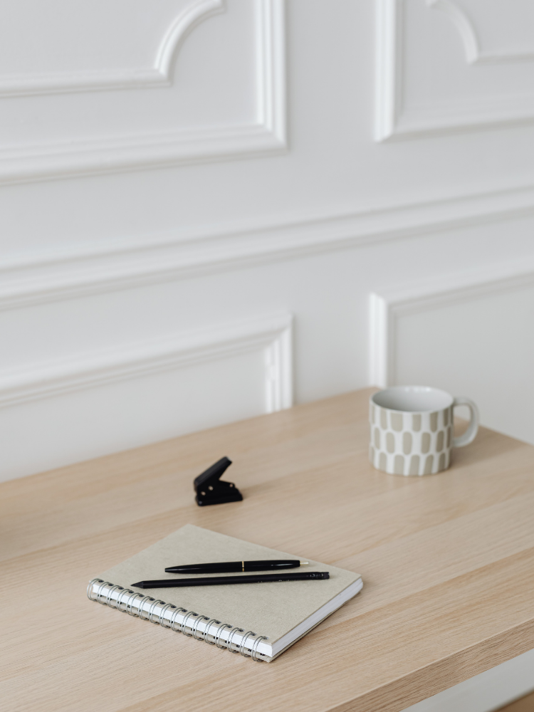 A wooden desk with a closed notebook, two black pens, a black stapler, a white mug with a beige geometric pattern, and a white panel wall in the background.