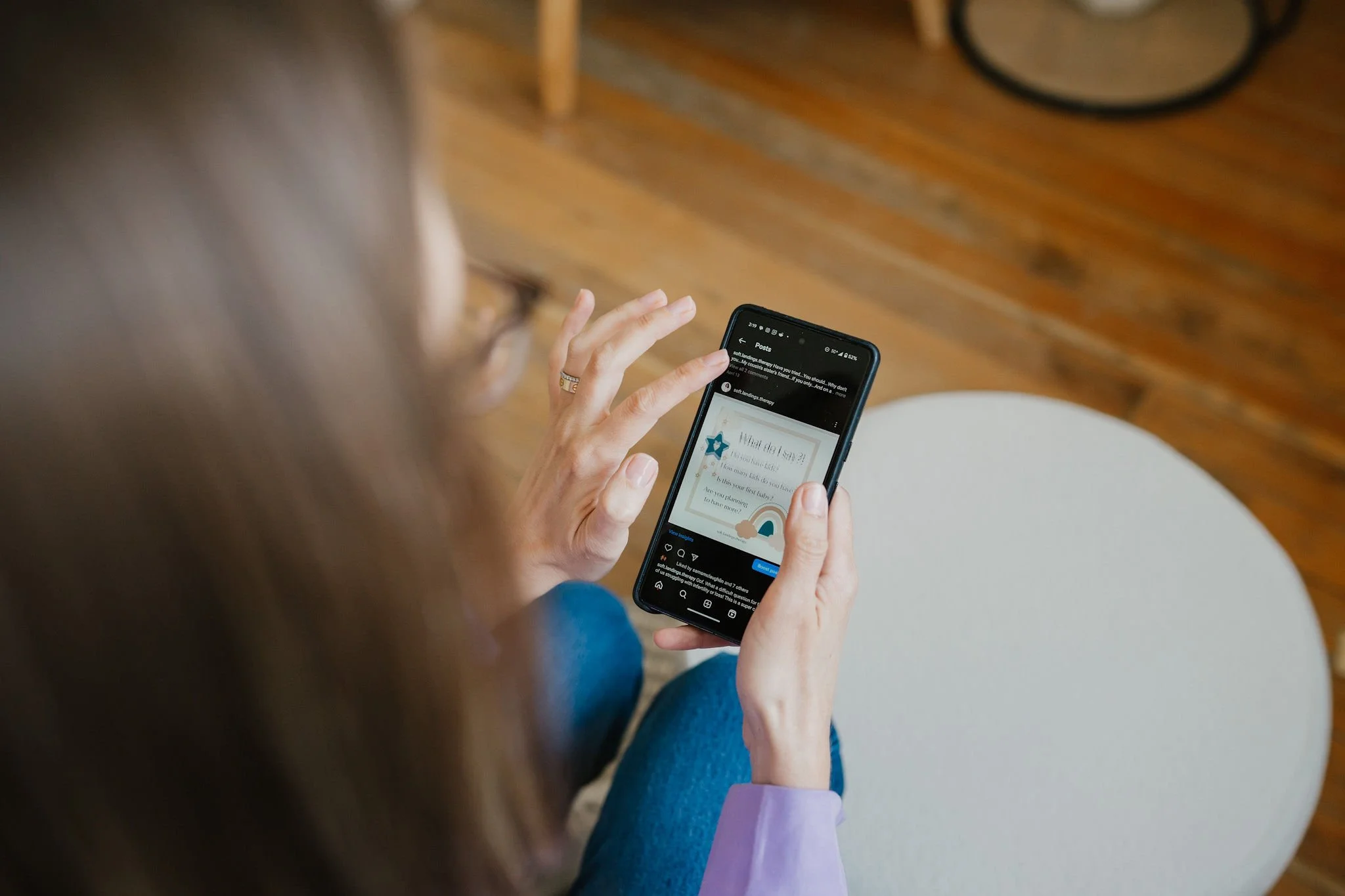 A person with long brown hair, wearing a purple long-sleeved shirt and blue jeans, is seated on a white chair, holding a smartphone. The person is using the phone, which displays a social media post, in a room with wooden floors, a wooden table leg, and a black circular item in the background.