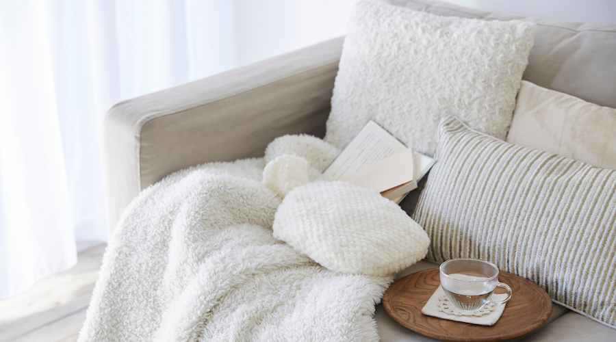 Cozy beige couch with fluffy white and striped pillows, an open book, a knit white blanket, and a wooden tray with a glass of tea.