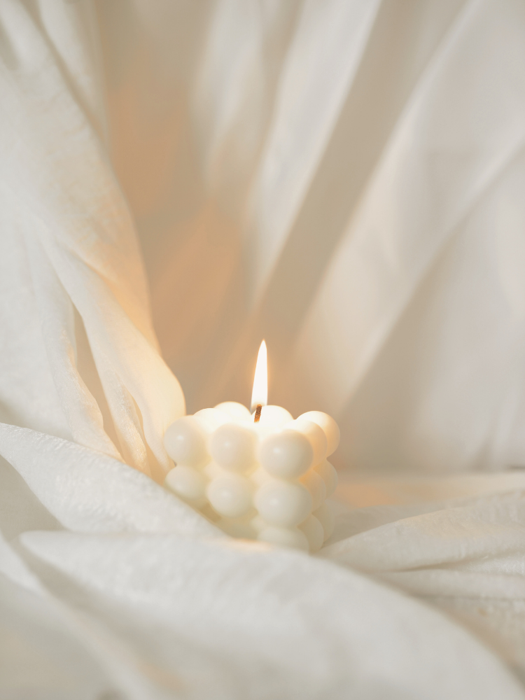 A white candle with a bubble-like design, lit, on soft white fabric, with a neutral background.