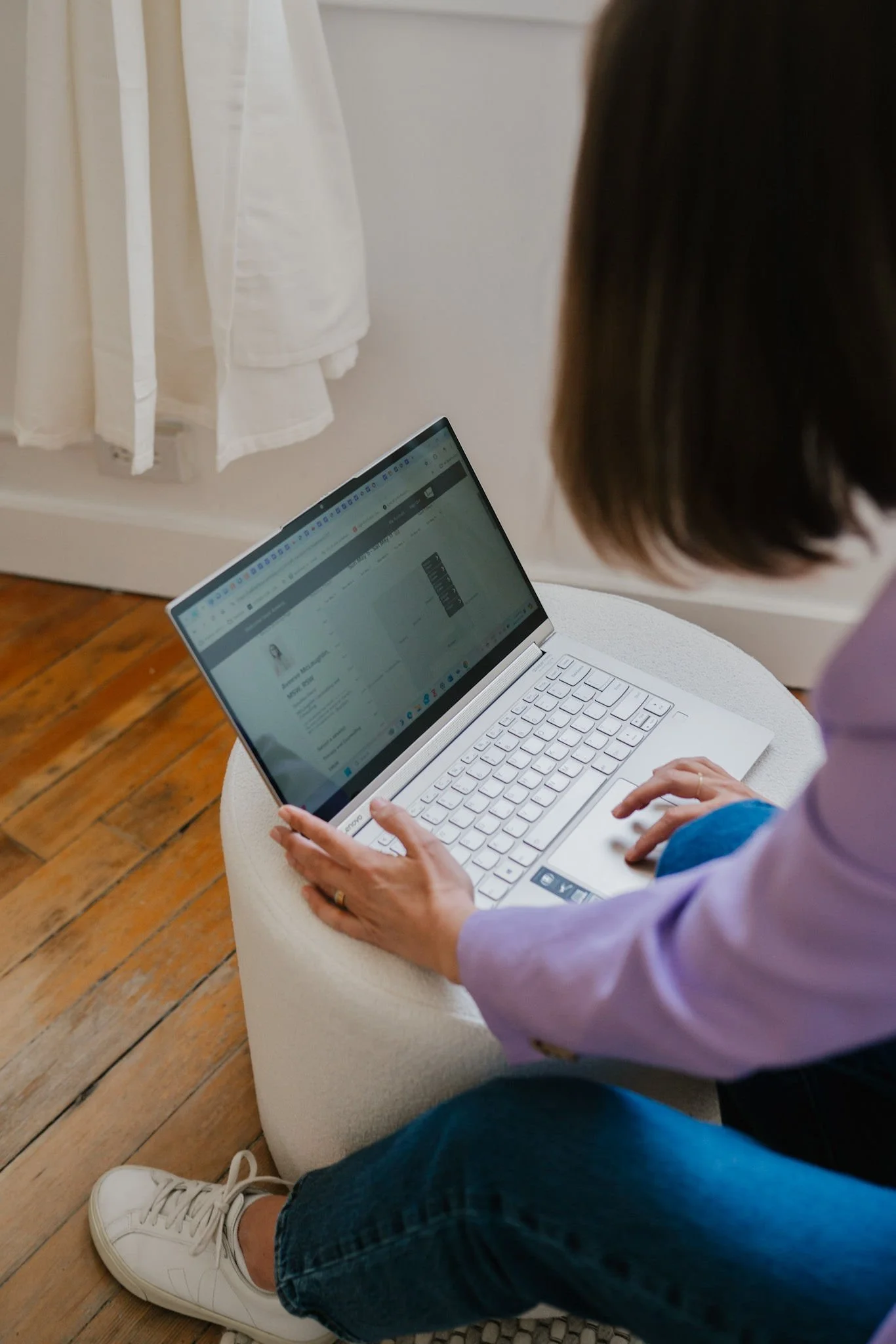 Person sitting on a white cushioned seat, wearing white sneakers, dark jeans, a purple jacket, and using a silver laptop on a round table. The floor is wooden, and there are white curtains hanging in the background.