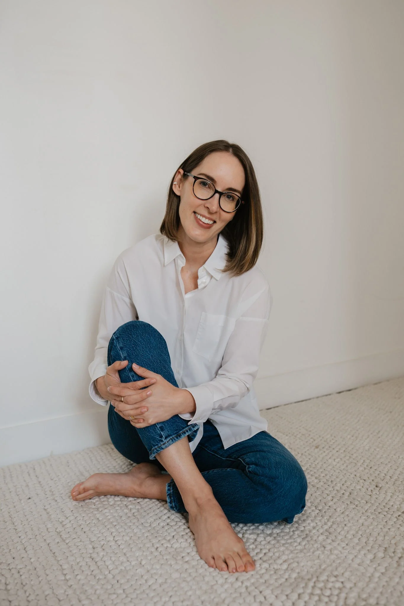 A woman with glasses and shoulder-length brown hair, wearing a white shirt and blue jeans, sitting barefoot on a beige textured carpet, smiling at the camera, with another person's legs in front of her.