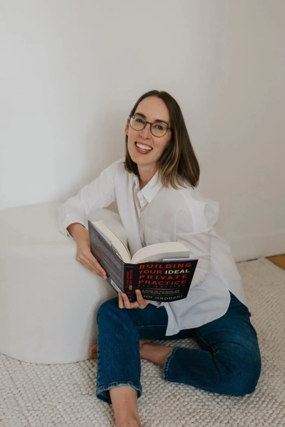 Aveeve McClaughlin | A woman with glasses and shoulder-length hair sitting on a beige textured rug, holding an open book titled "Building Your Ideal Private Practice". She is smiling, wearing a white button-up shirt and blue jeans.