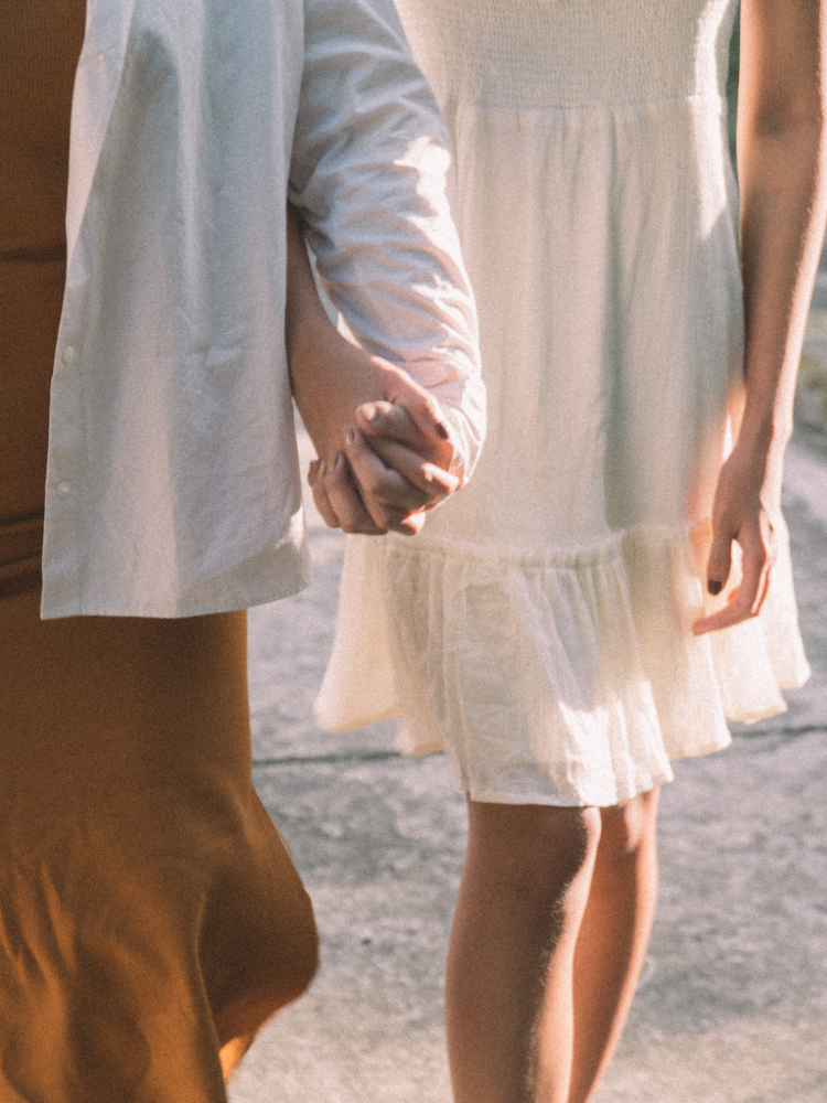Close-up of a couple holding hands, with one wearing a light-colored dress and the other wearing a white shirt, on a sunny day.